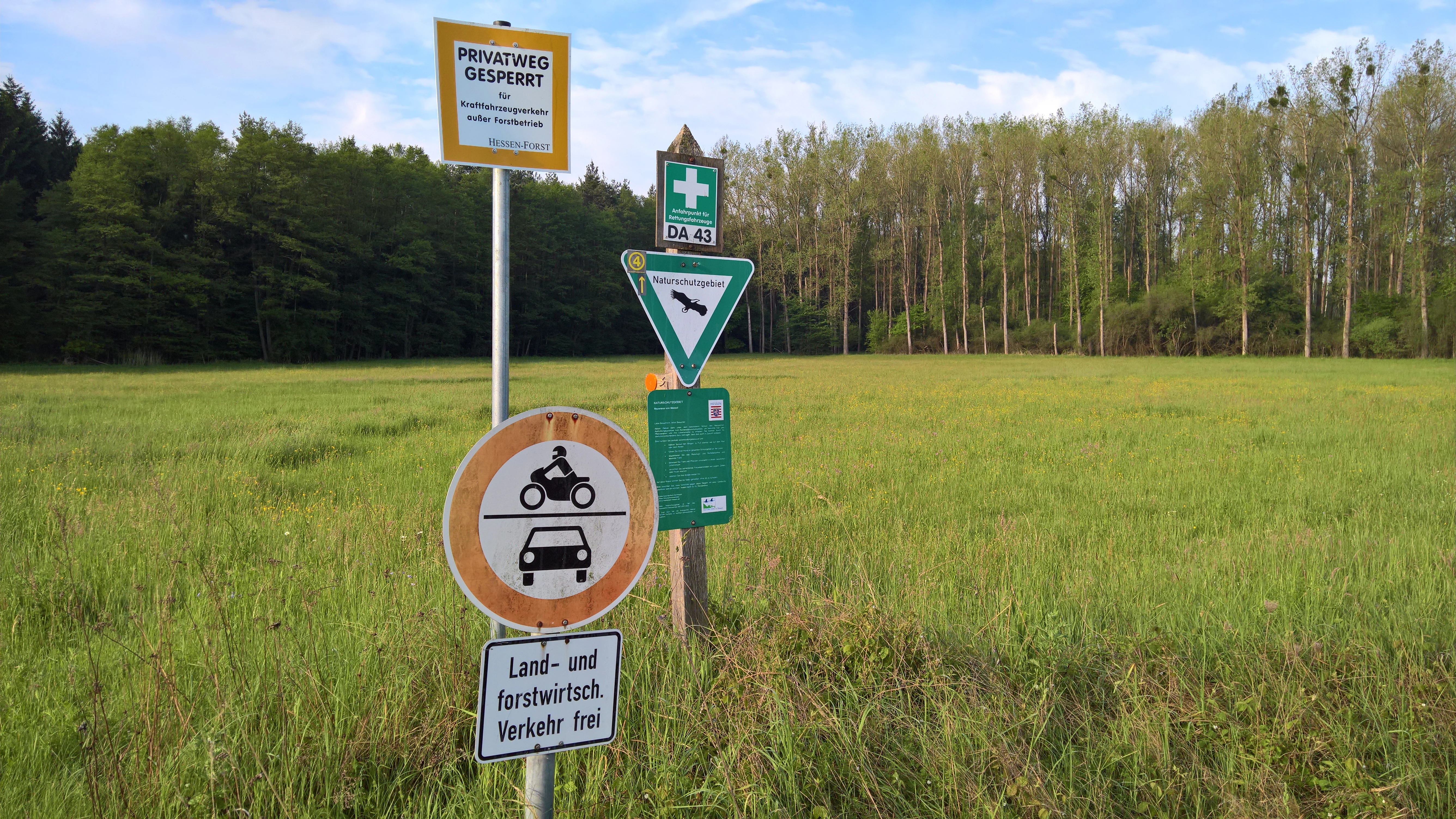 A group of traffic and nature reserve signs standing on a metal pole in front of a green meadow and a line of trees.