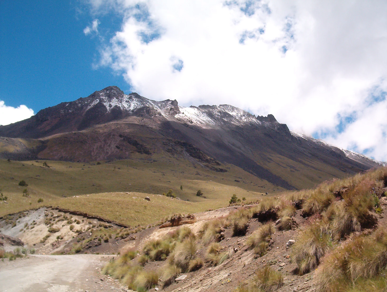 A wide-angle mountain landscape shows a large volcanic peak under a partially cloudy blue sky; the upper slopes are dusted with snow, while the foreground consists of dry, grassy volcanic terrain.