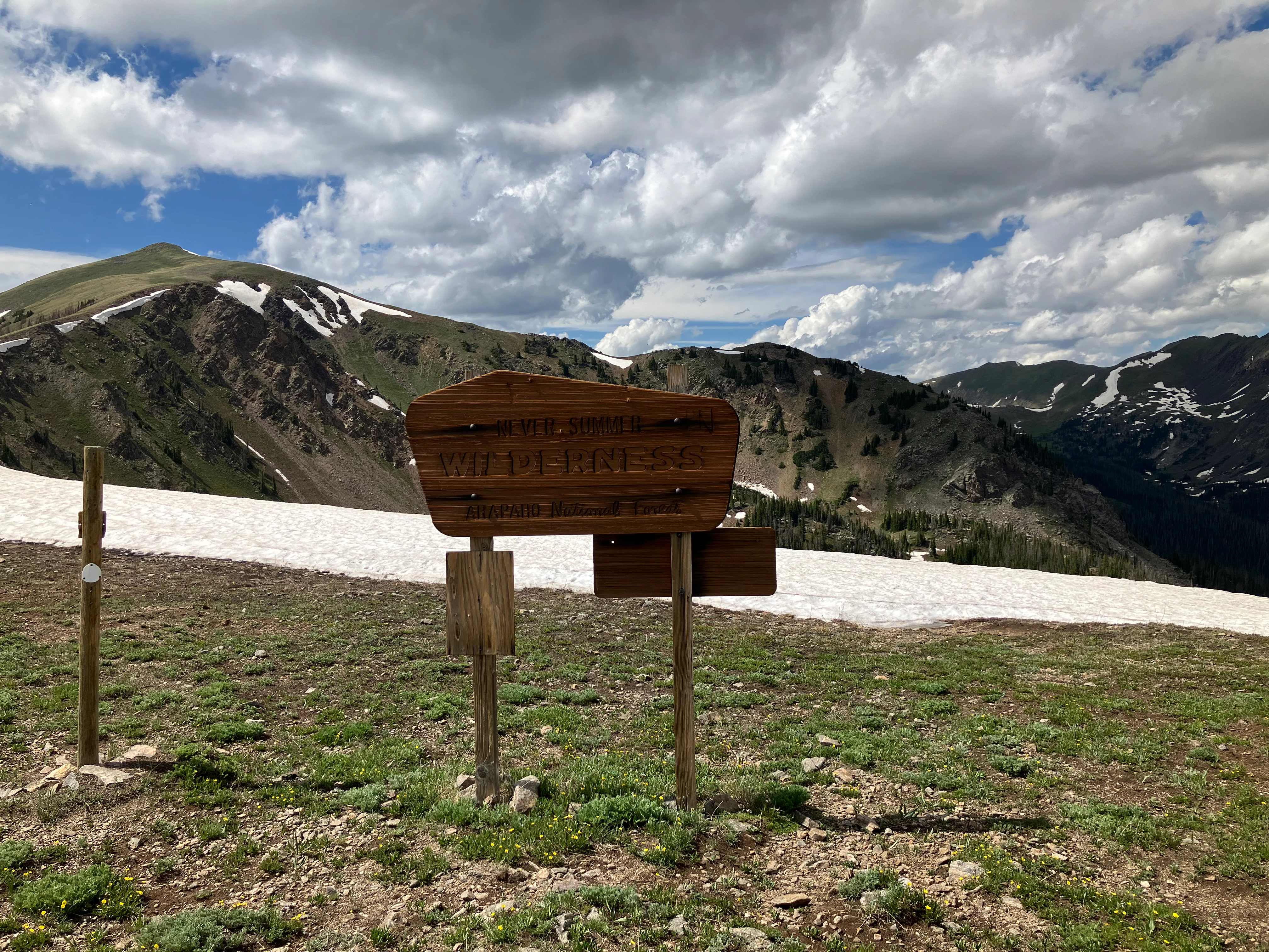 A weathered brown wooden sign marked Never Summer Wilderness stands in an alpine meadow with patches of snow and rocky peaks in the background.