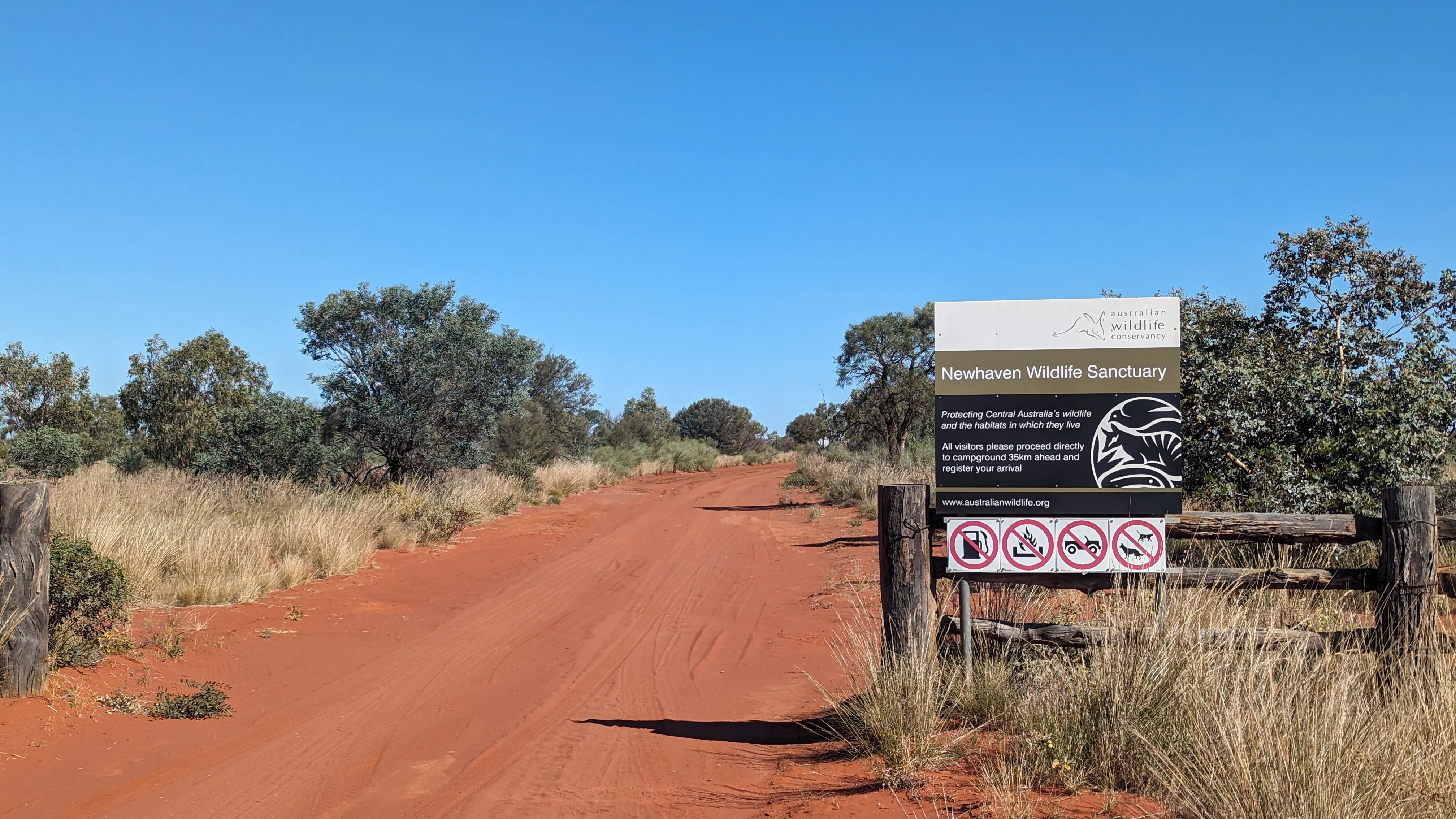 A sign for Newhaven Wildlife Sanctuary mounted on a wooden fence next to a red dirt road surrounded by arid shrubs.