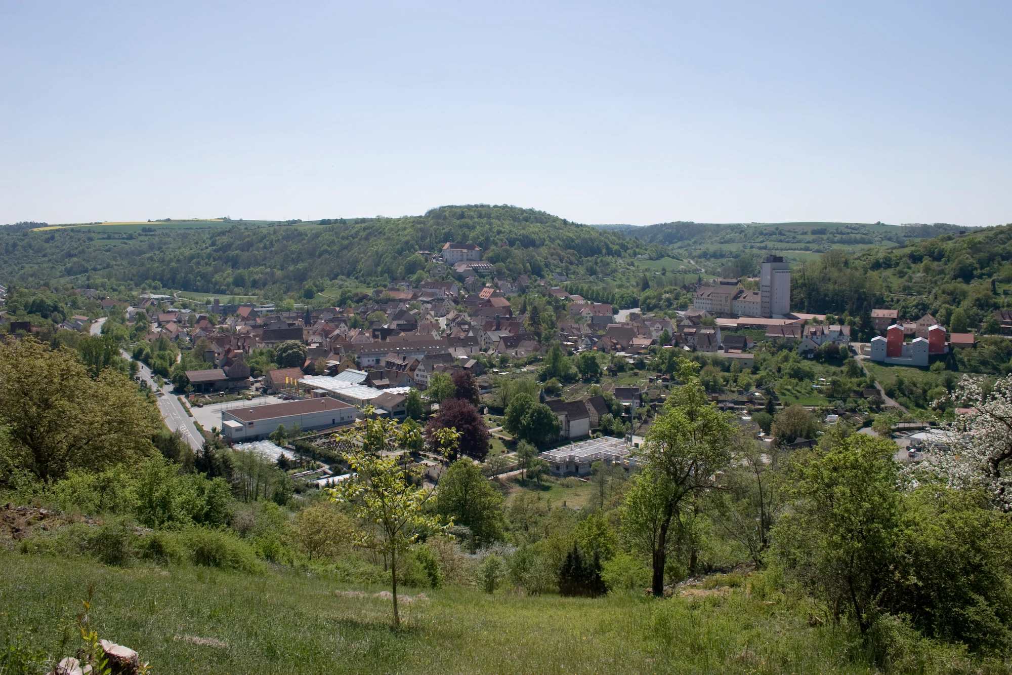 Vista panorámica del municipio de Niederstetten en un valle, con colinas boscosas al fondo y vegetación verde en primer plano bajo un cielo despejado.