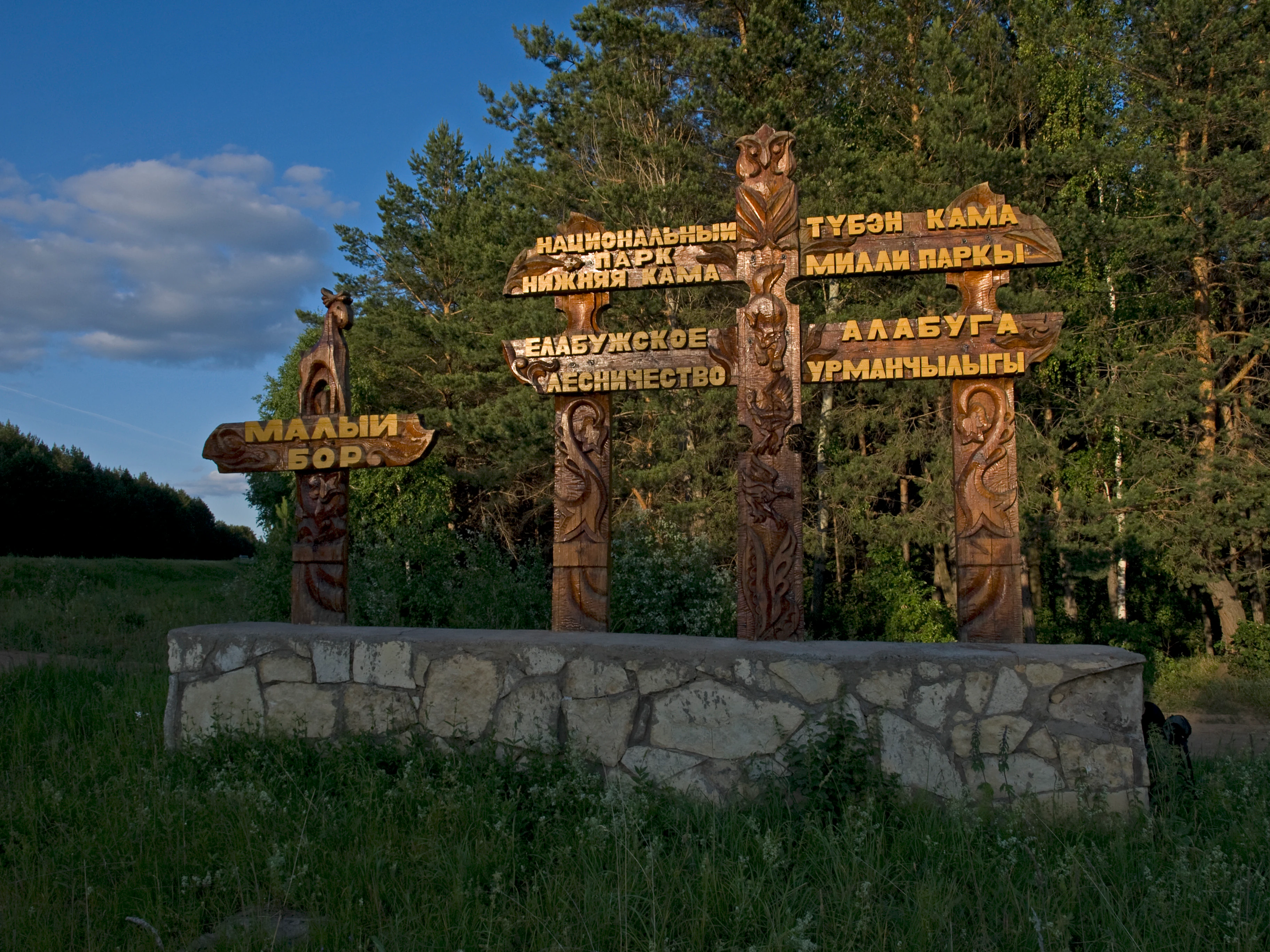 A rustic, hand-carved wooden sign with Cyrillic text stands on a stone base in front of a forest, marking the entrance to the Maly Bor section of a national park.