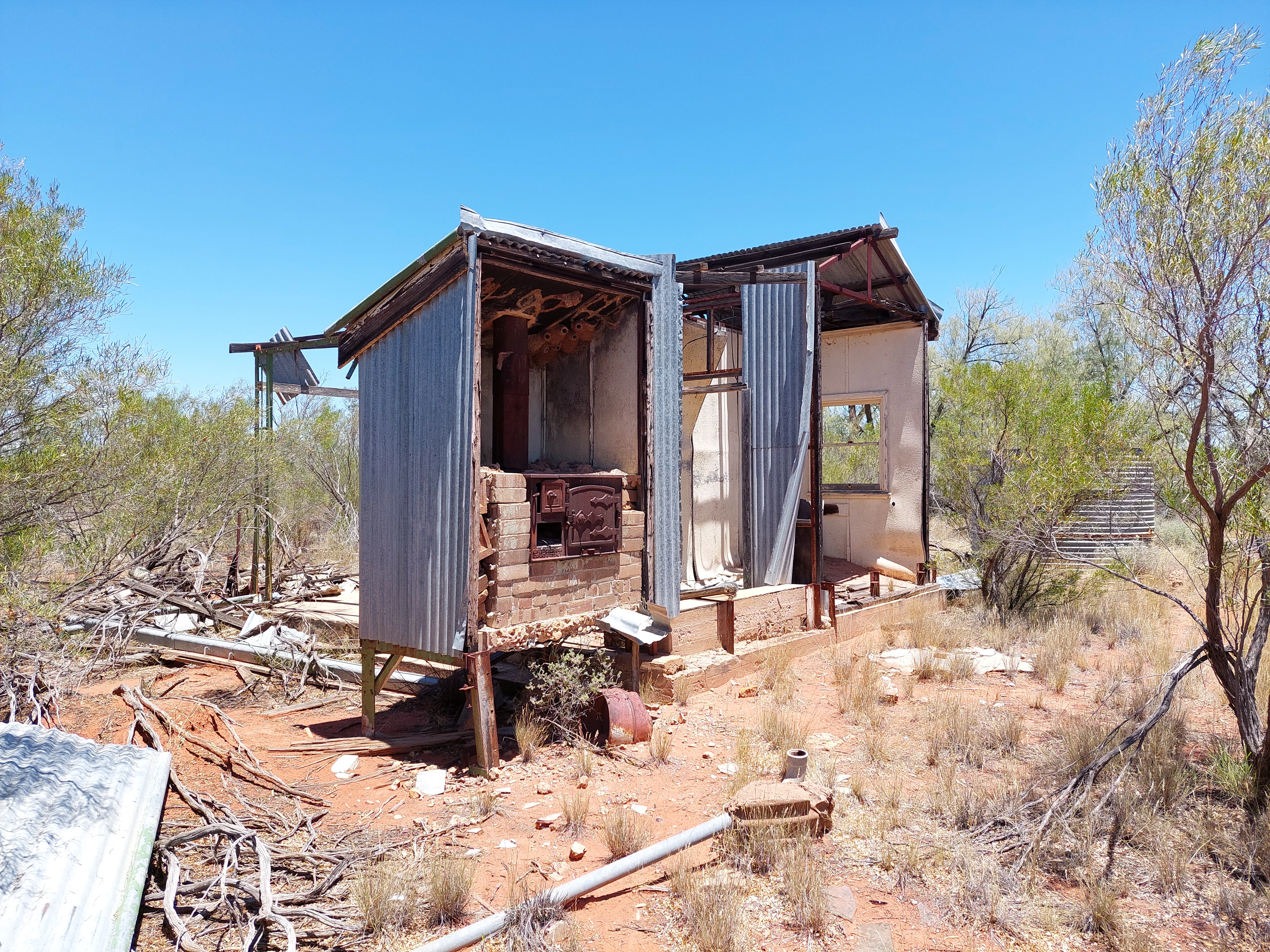 Remains of a small, rustic shed with corrugated metal walls and a brick fireplace, situated in an arid, scrubby environment.