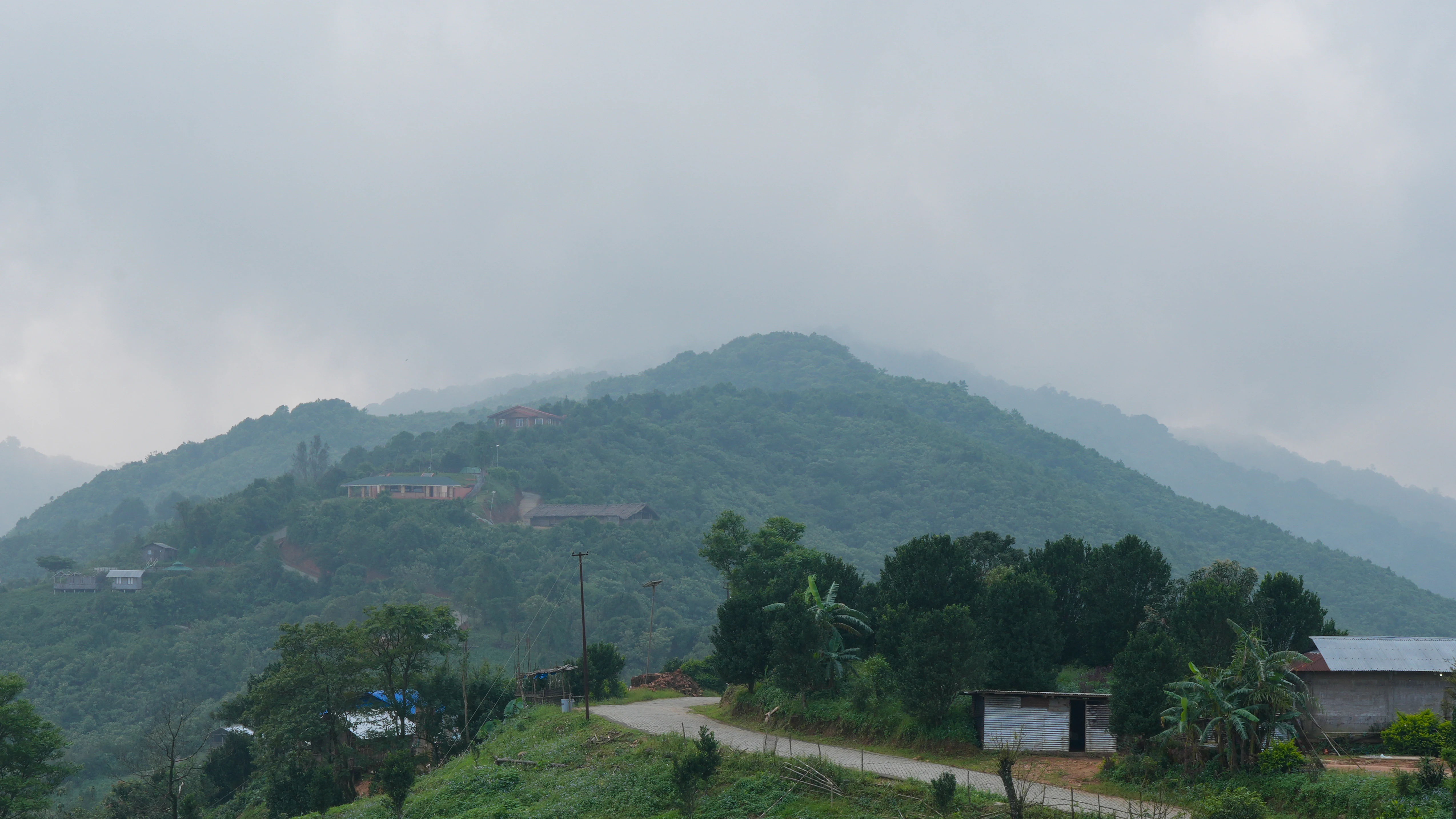 Lush green mountain ridge under cloudy sky with road and small buildings in foreground