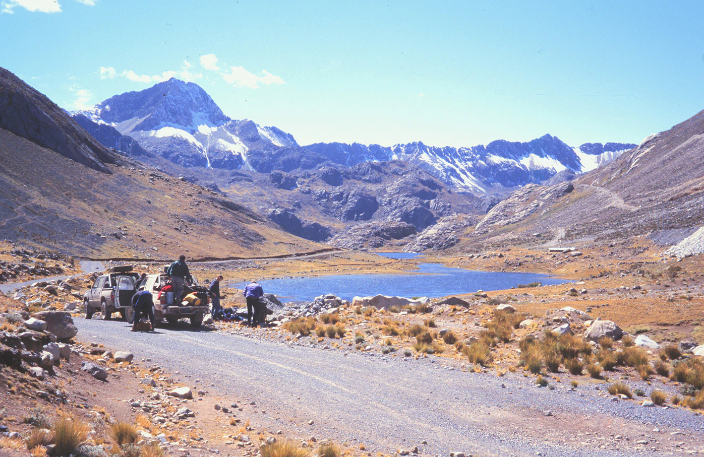Una carretera de grava en primer plano con una camioneta estacionada y personas cerca de ella, que conduce hacia un pequeño lago azul enclavado en un valle rodeado de montañas rocosas y nevadas bajo un cielo azul despejado.