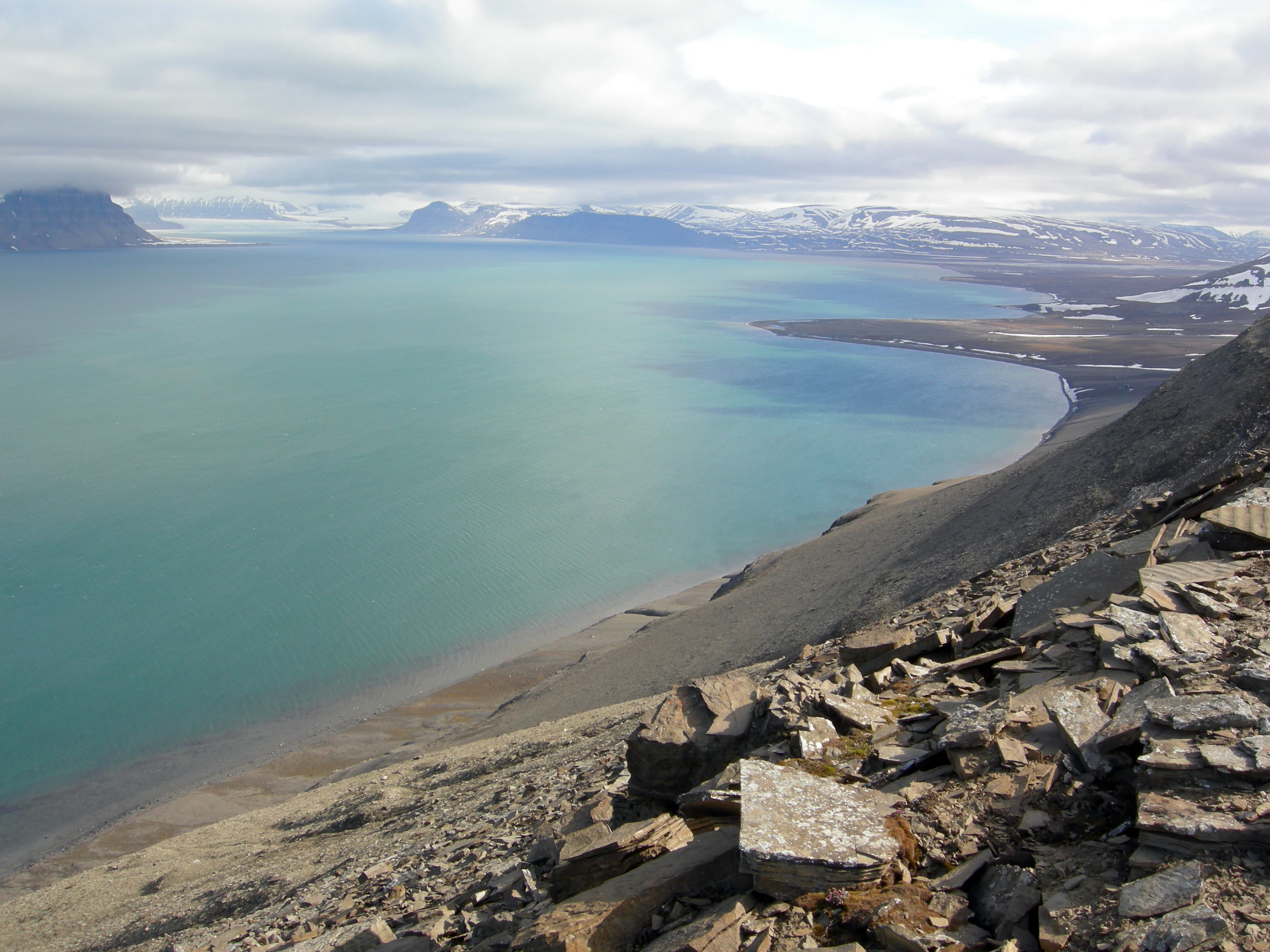 Wide view of turquoise fjord water, rocky shoreline, and snow-capped mountains under cloudy sky