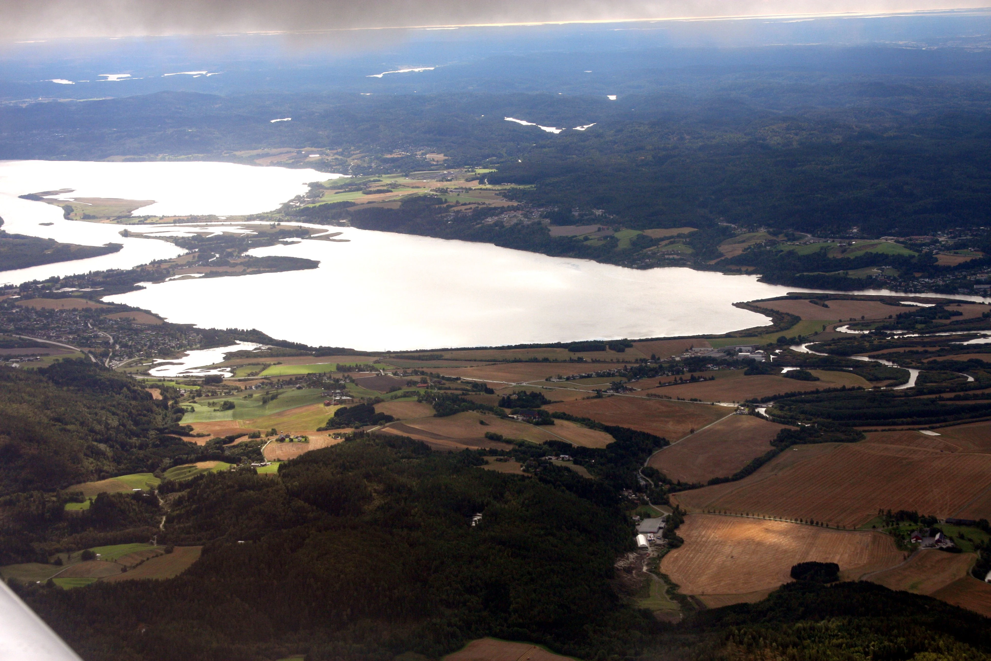 Aerial photograph showing a large lake and branching wetland delta surrounded by forest patches and agricultural fields, with distant tree-covered hills on the horizon.