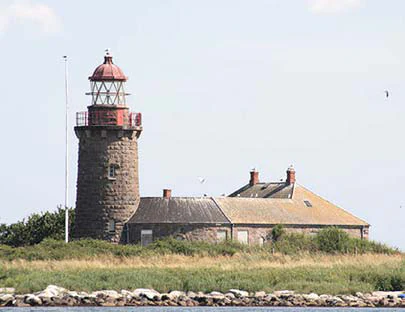 A historic stone lighthouse with a red metal lantern room stands next to low buildings on a grassy island in the Kattegat sea.