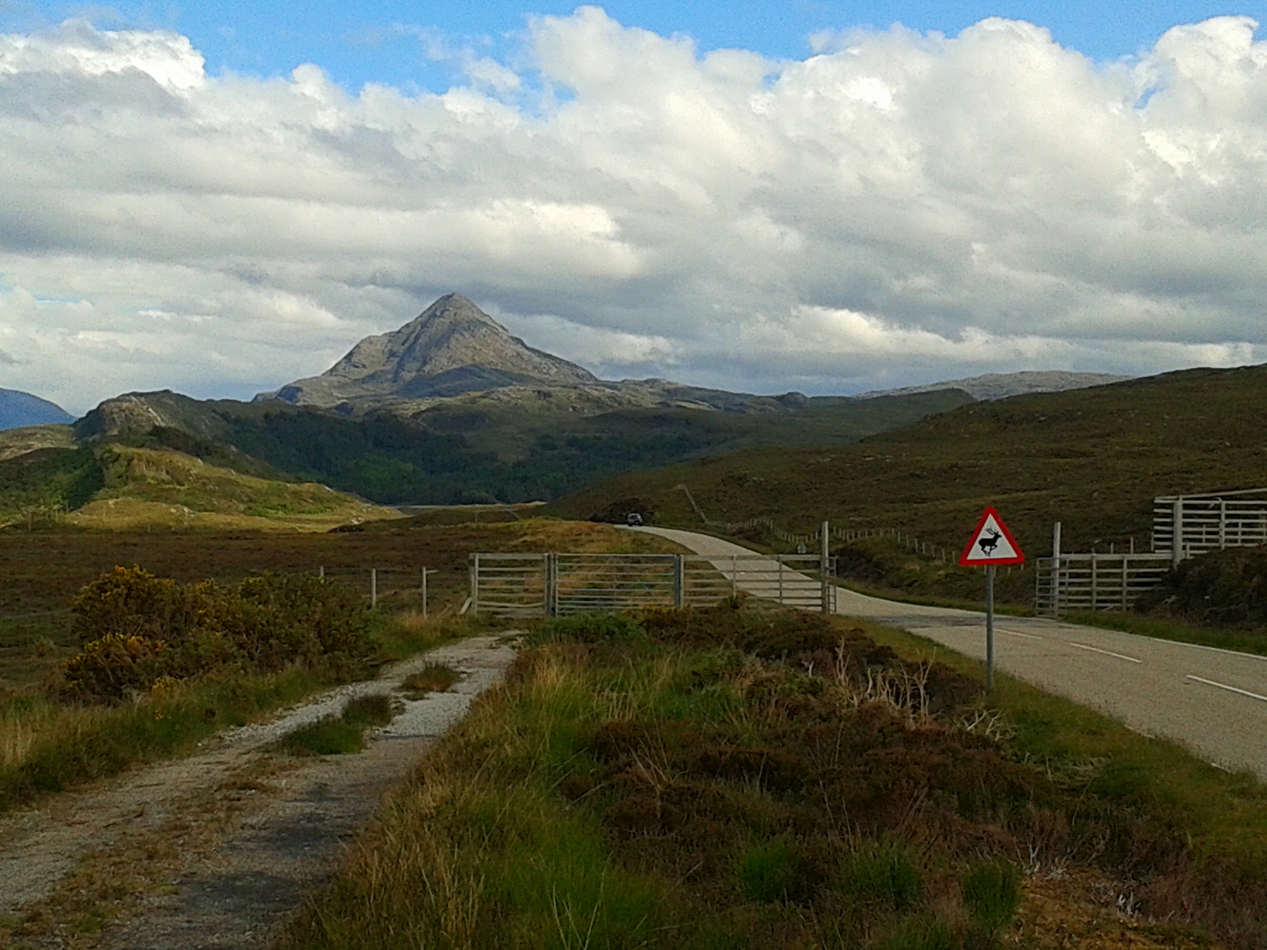 A conical mountain peak rises in the distance behind rolling moorland, with a paved road, metal cattle grid gate, and a triangular warning sign in the foreground under a partly cloudy sky