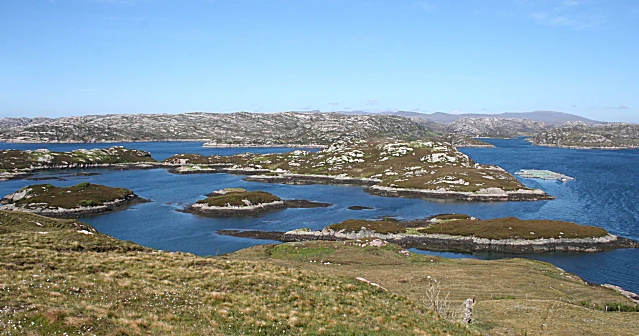 A wide landscape view of a blue loch dotted with small, rocky, grass-covered islands under a clear blue sky, seen from a grassy hillside.