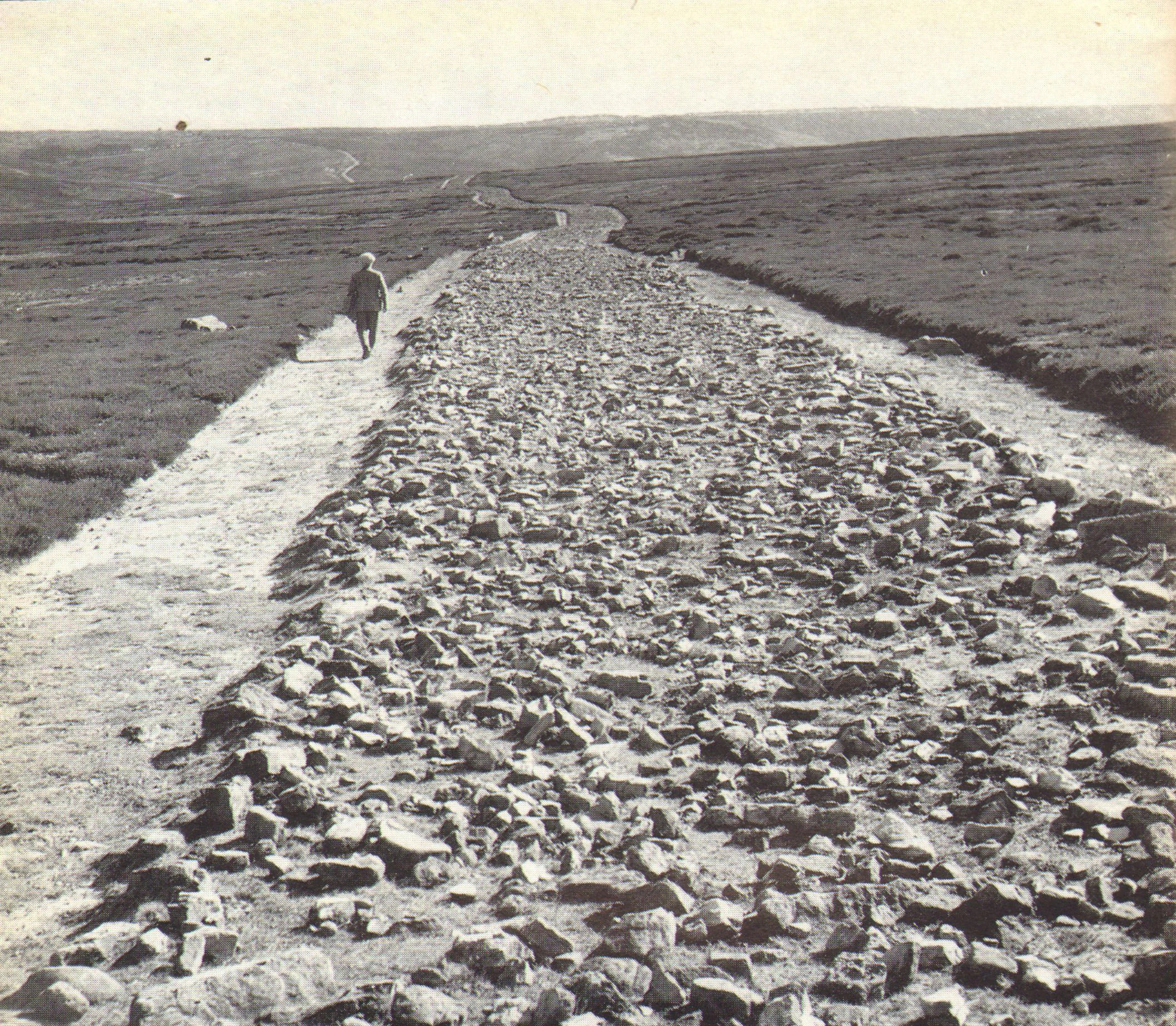 Black and white photograph showing a long stone road stretching across moorland with a person walking on the left side