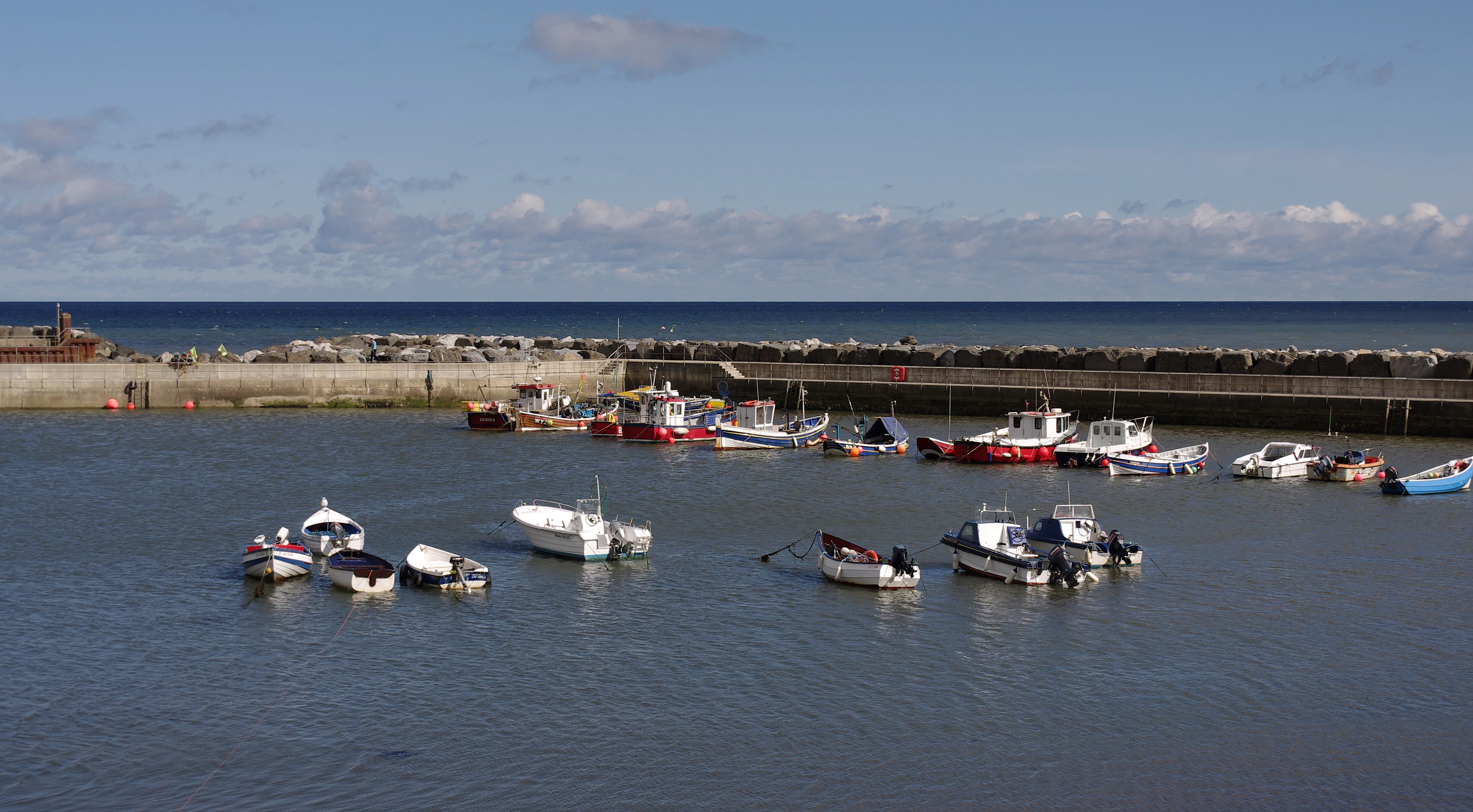 Several small boats floating on calm water in a harbour with stone breakwater and distant sea under a partly cloudy sky