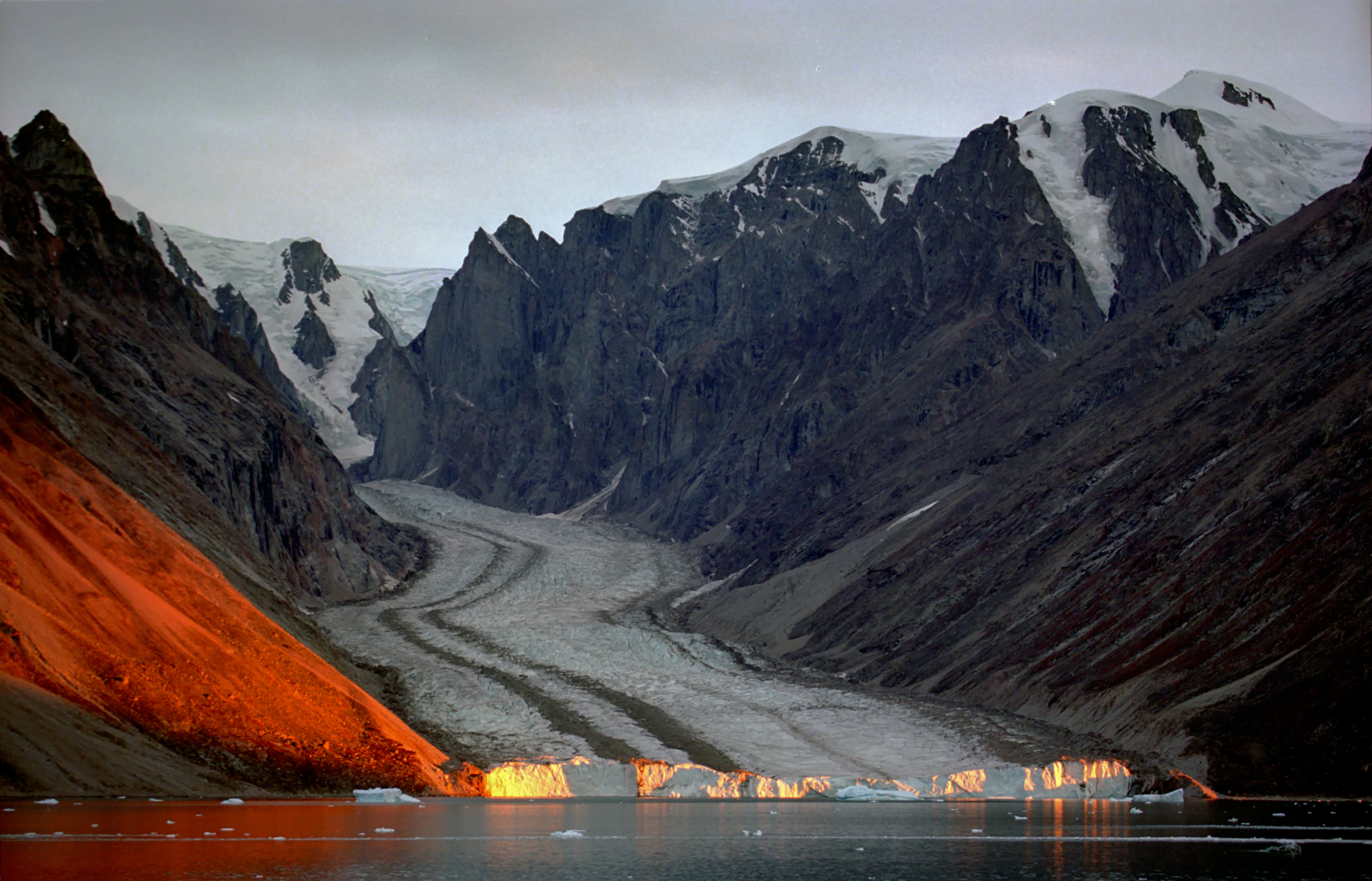 A glacier in Franz Josef Fjord surrounded by snow-capped mountains and orange-tinted ice reflecting light