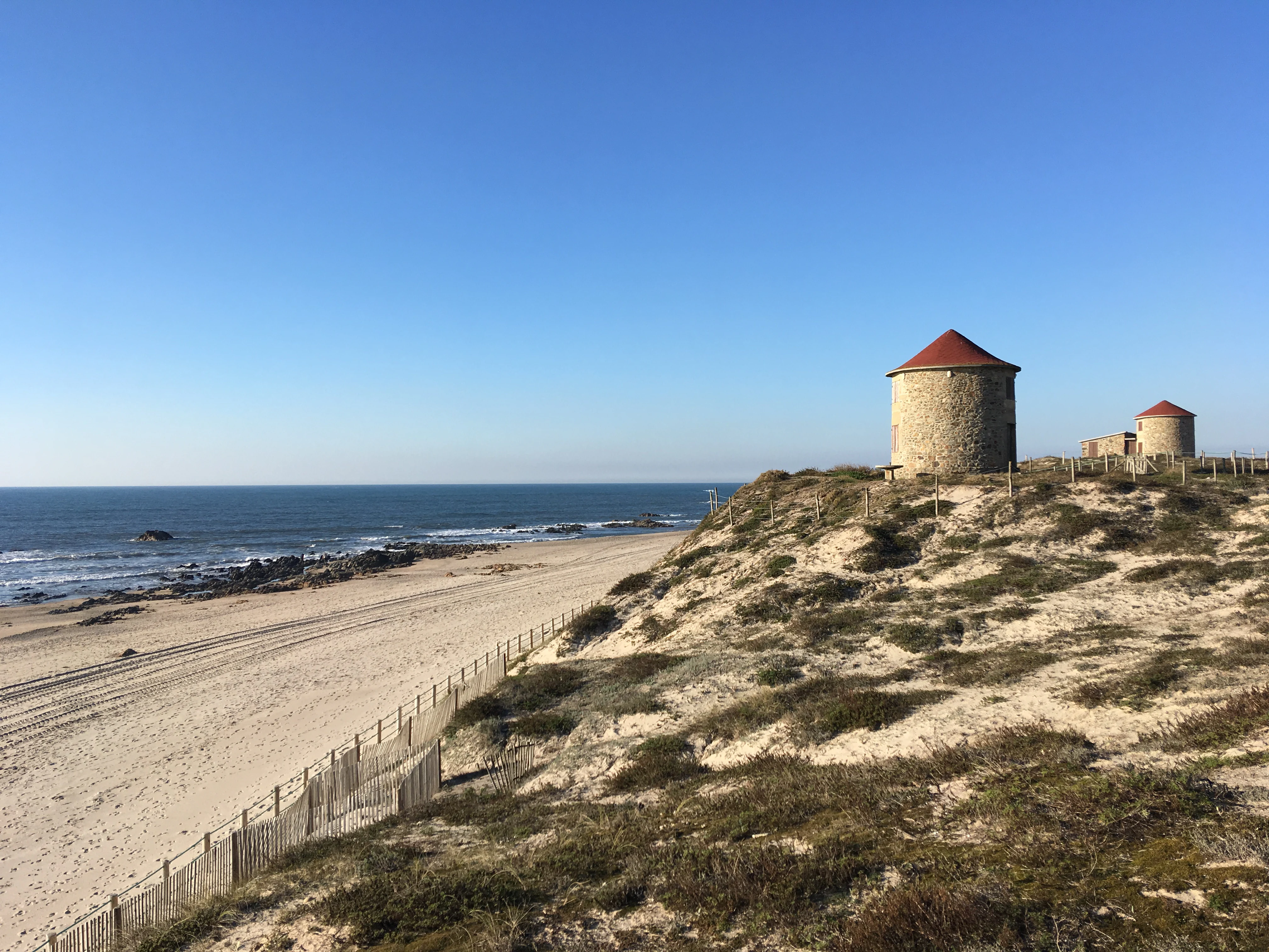 Sand dunes with two windmills featuring red roofs, a wooden fence, and a sandy beach leading to the ocean under a clear blue sky