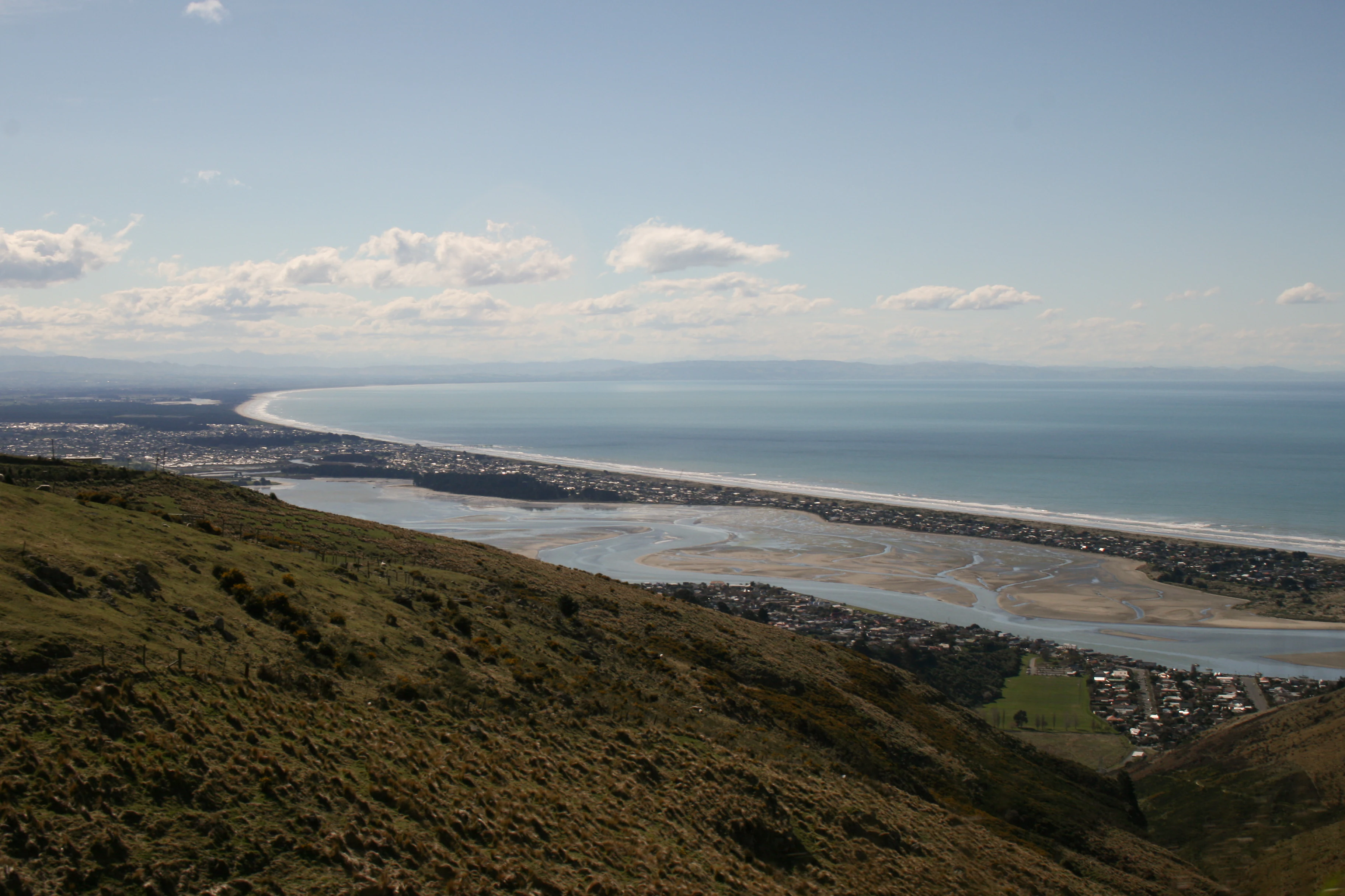 A wide, high-angle landscape shot looking down from a grassy hillside onto a long sandy coastline, an estuary with channels, and the ocean under a clear sky.