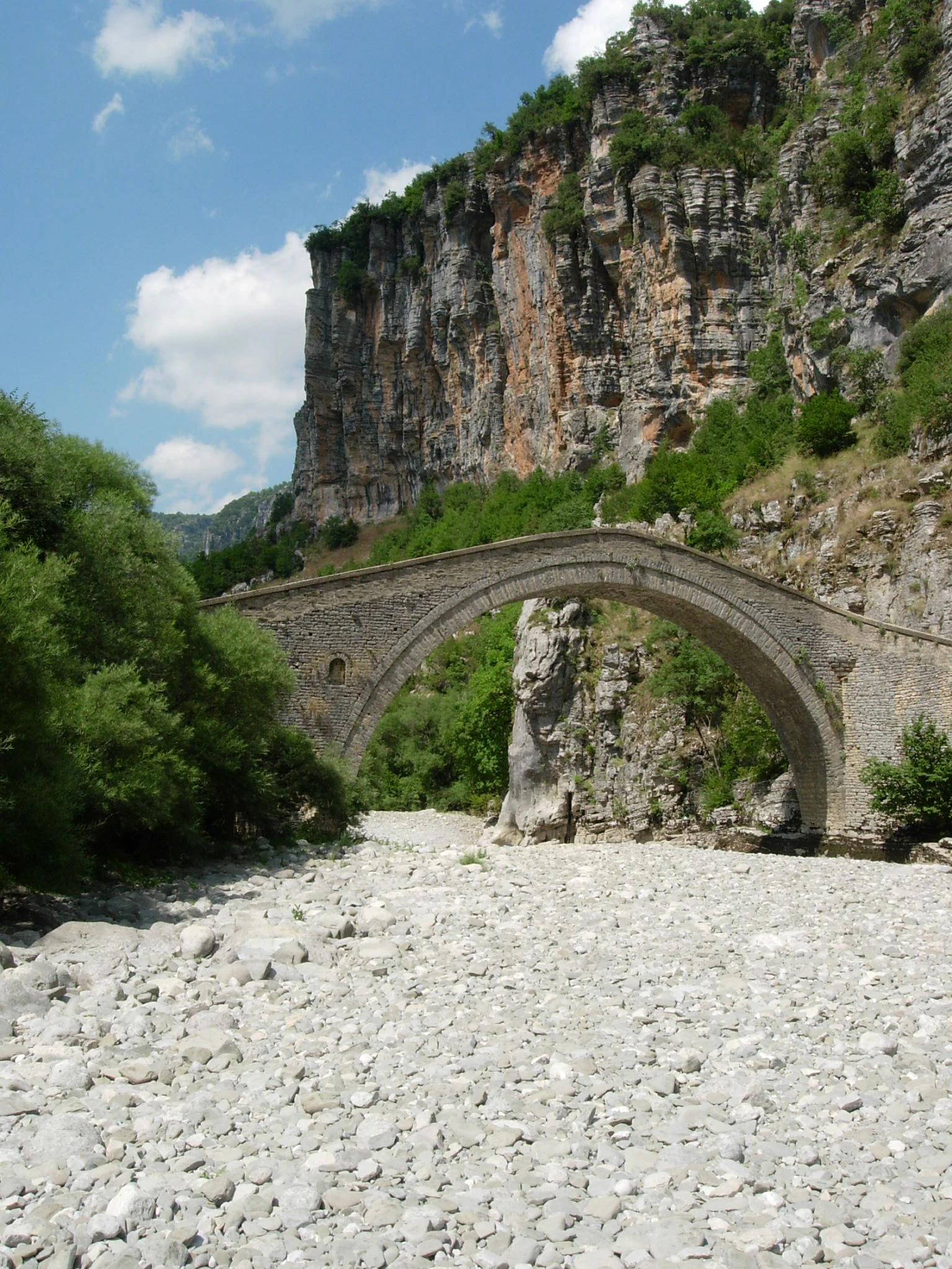 Un histórico puente de piedra arqueado se alza en un seco lecho rocoso de río, al pie de un imponente acantilado de caliza escarpada.