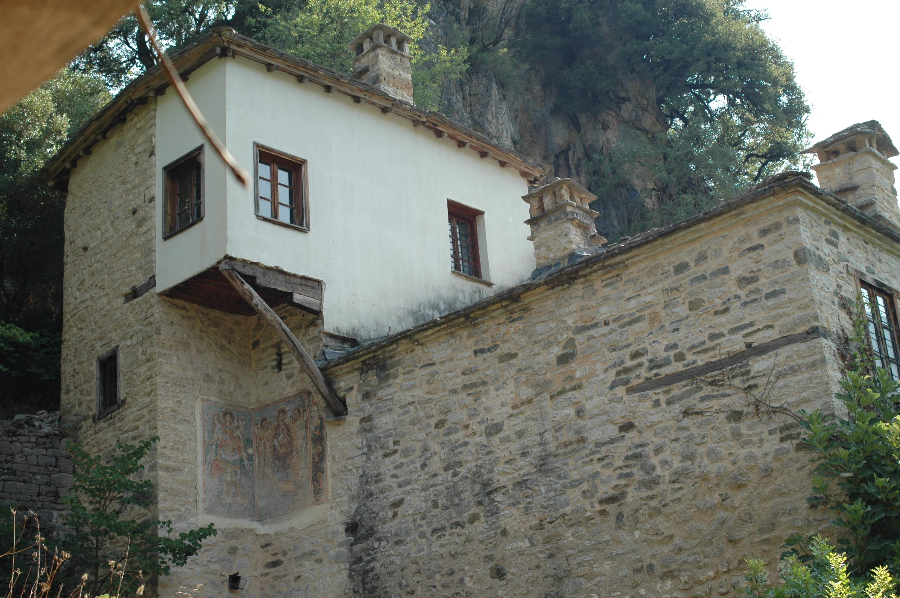 Un edificio monástico de piedra y yeso de varias plantas construido contra un acantilado rocoso, con mampostería tradicional y un fresco exterior en la pared.