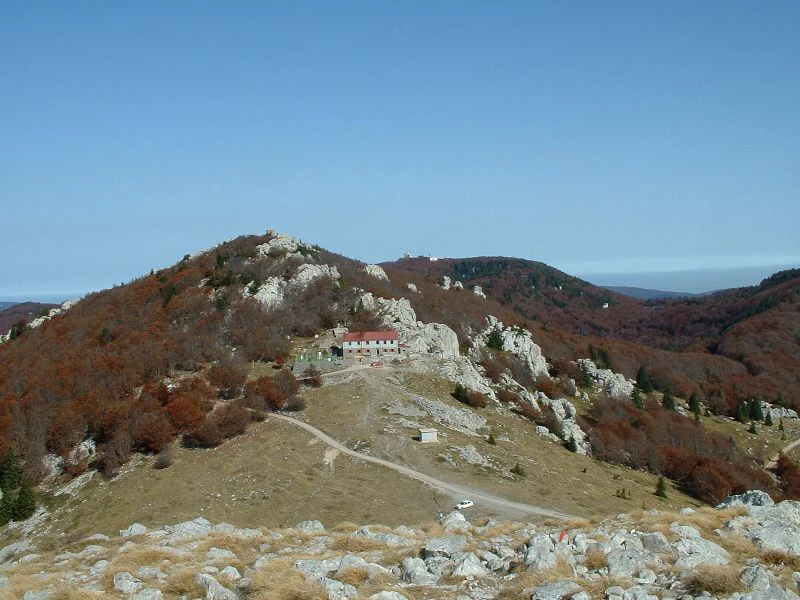 Rocky hill with a small building featuring a red roof, surrounded by brown and orange foliage on a clear day with distant mountain ranges visible.