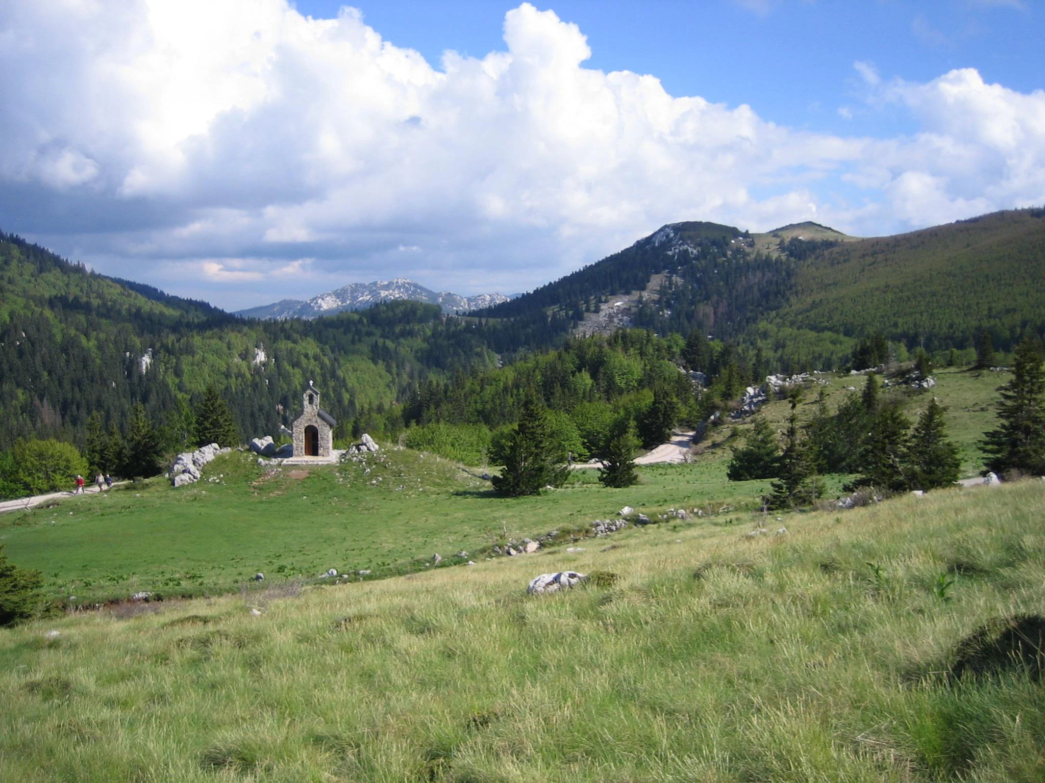 Grassy hillside with scattered rocks, a small stone chapel, and forested mountains under a partly cloudy sky