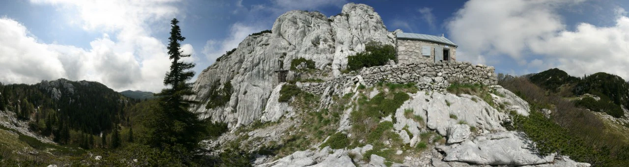 Stone hut atop rocky hillside with mountain peaks and forested valley under partly cloudy sky