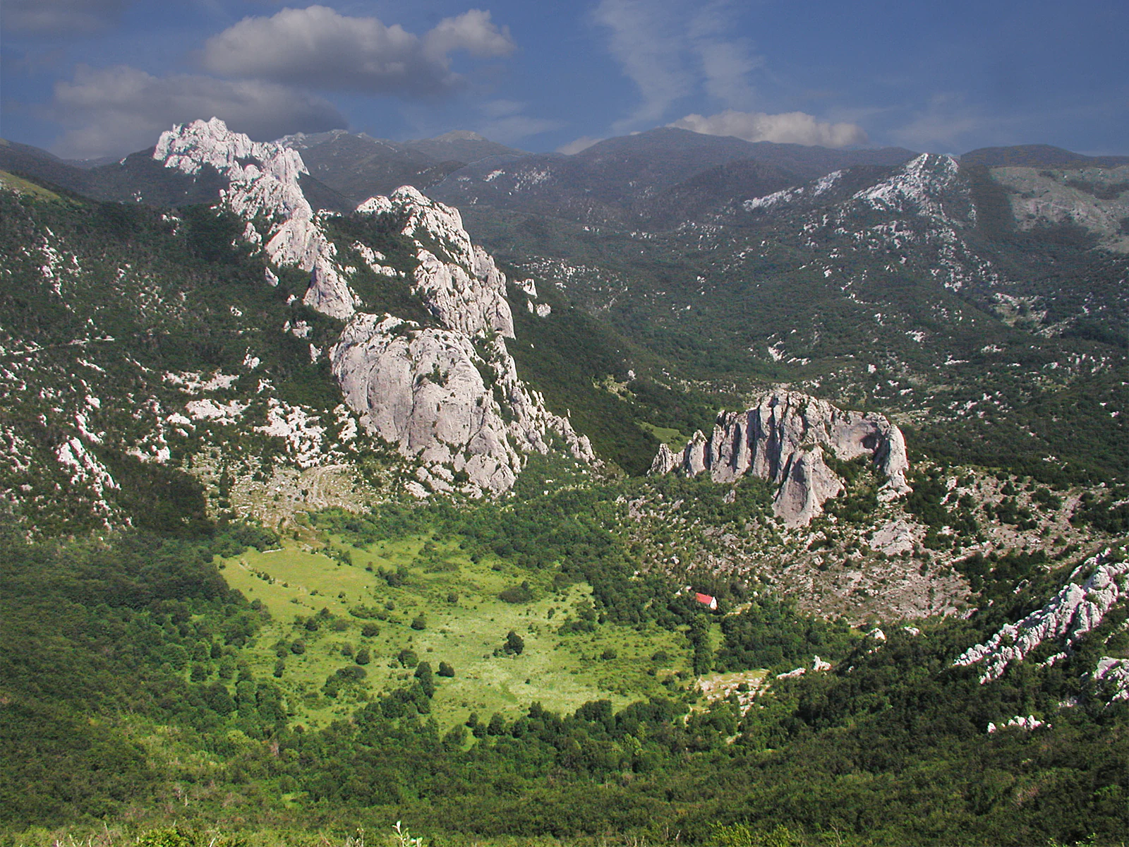 Wide view of rocky mountain peaks with green valleys and scattered vegetation under a partly cloudy sky.