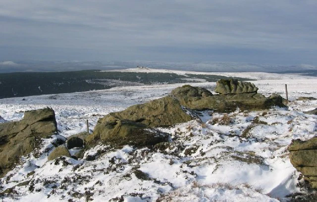 Rocky summit with snow-covered ground and distant moorland under a partly cloudy sky