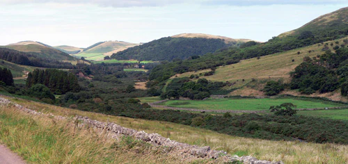 Panoramic view of a valley with rolling hills, stone walls, grassy fields, and distant mountains under a partly cloudy sky