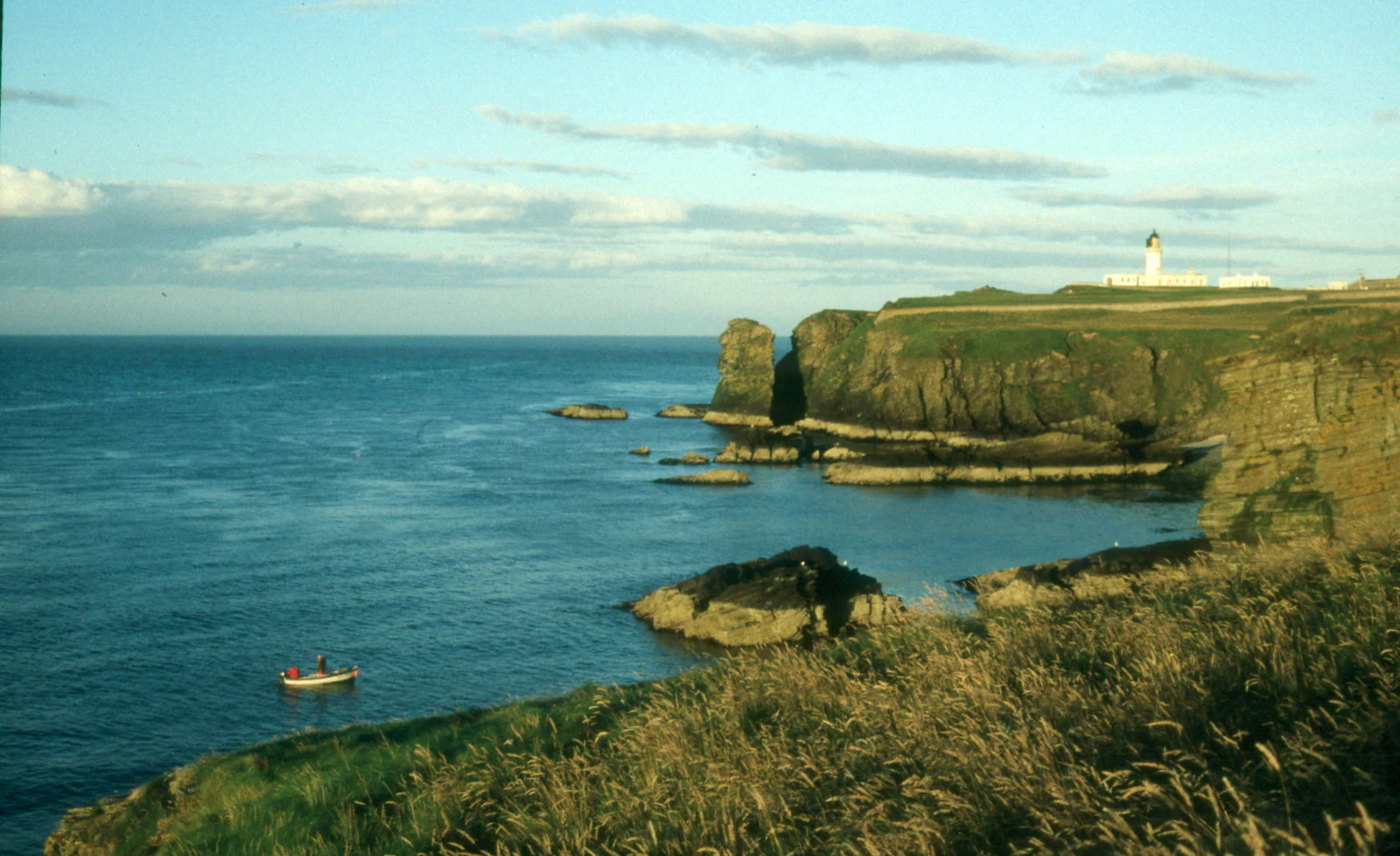 Coastal view at Noss Head showing sea cliffs, a lighthouse in the distance, and a small boat on the water.