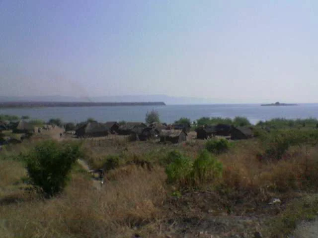 Paisaje seco y herbáceo con arbustos dispersos, una aldea con estructuras sencillas y el lago Tanganica al fondo bajo un cielo brumoso