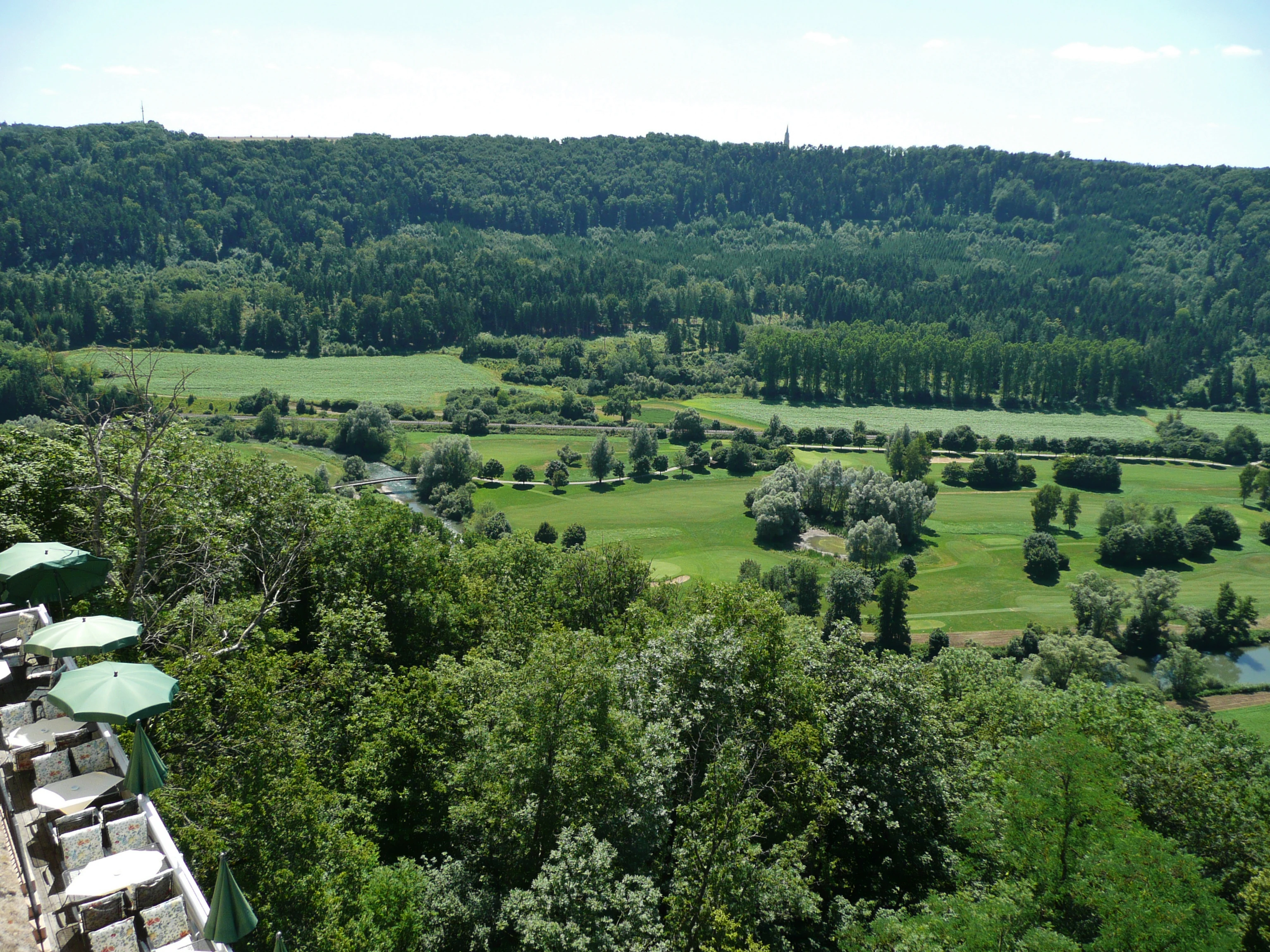Vista en ángulo elevado de un valle verde y frondoso con prados, bosques y un río serpenteante, visto desde una terraza con mesas de restaurante al aire libre y sombrillas verdes en primer plano.