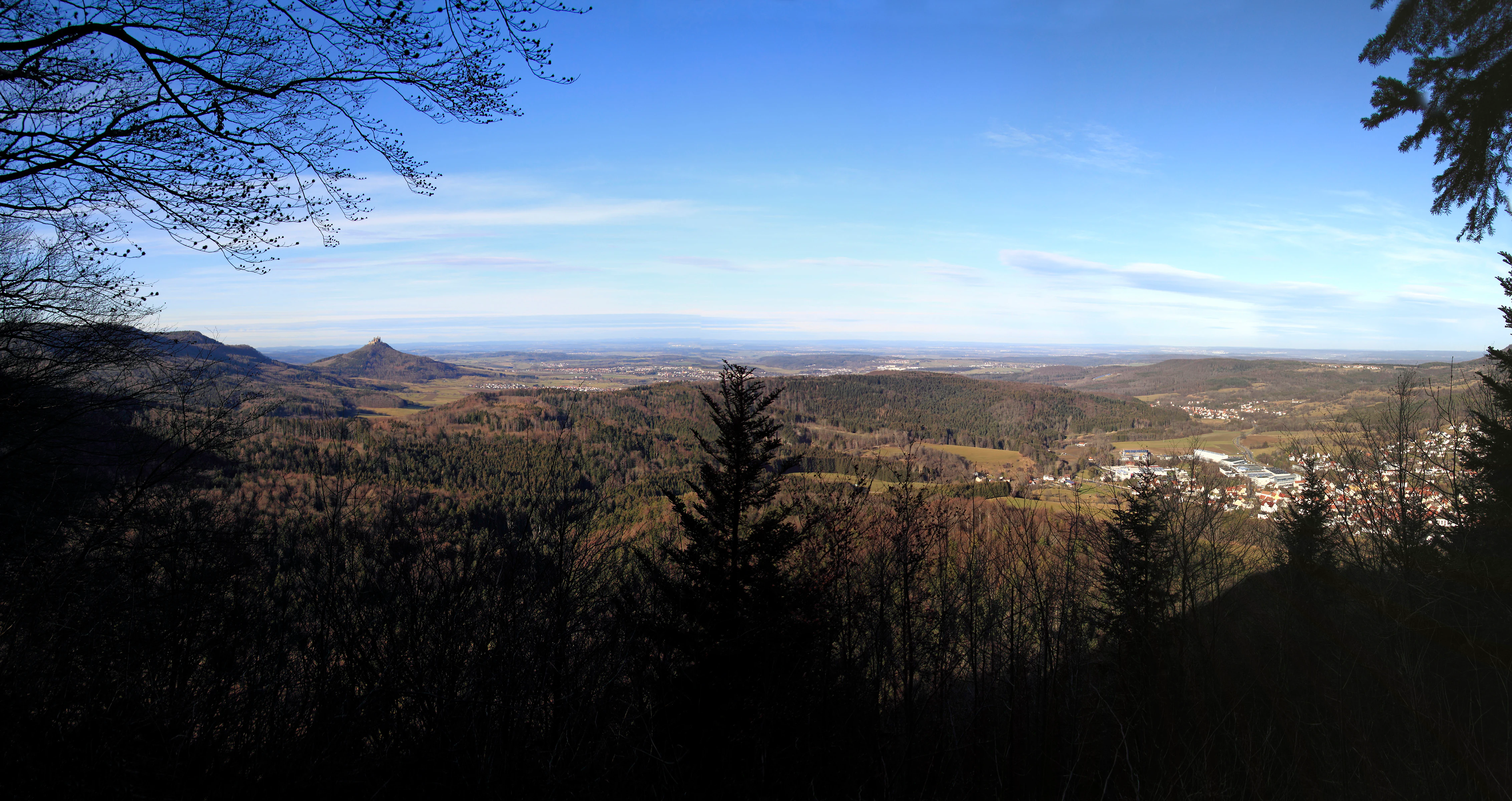 Panoramic landscape view from a high vantage point, looking out over a valley with rolling hills, dense forests, and a distant town, under a clear blue sky.