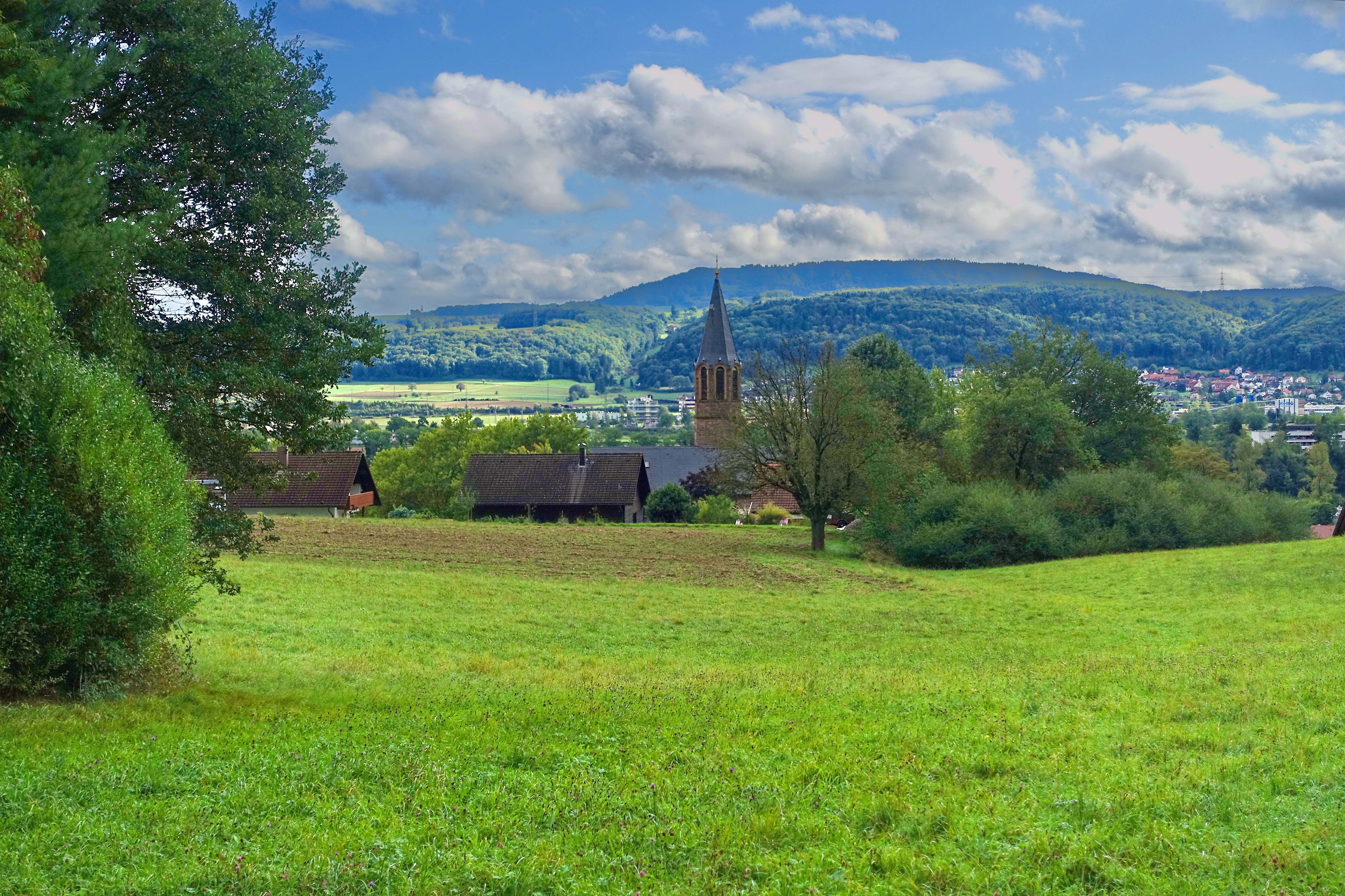 A green, rolling meadow in the foreground leads to a small group of farmhouses and a tall, slender church tower; rolling, tree-covered mountains rise in the background under a blue sky with scattered clouds.