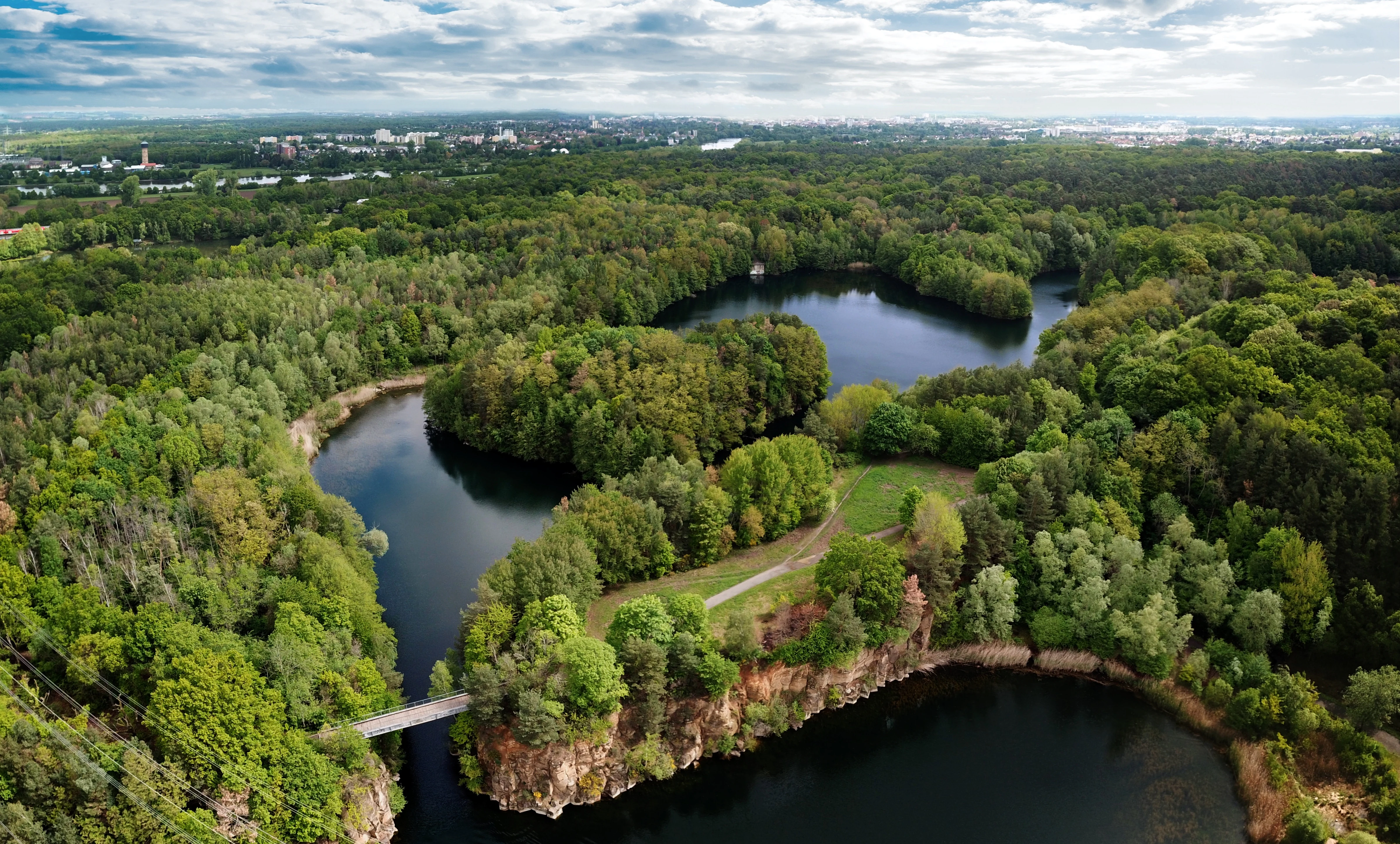 Aerial view of a nature reserve featuring dark blue lakes separated by forested land and rocky cliffs, with a pedestrian bridge connecting a landmass.
