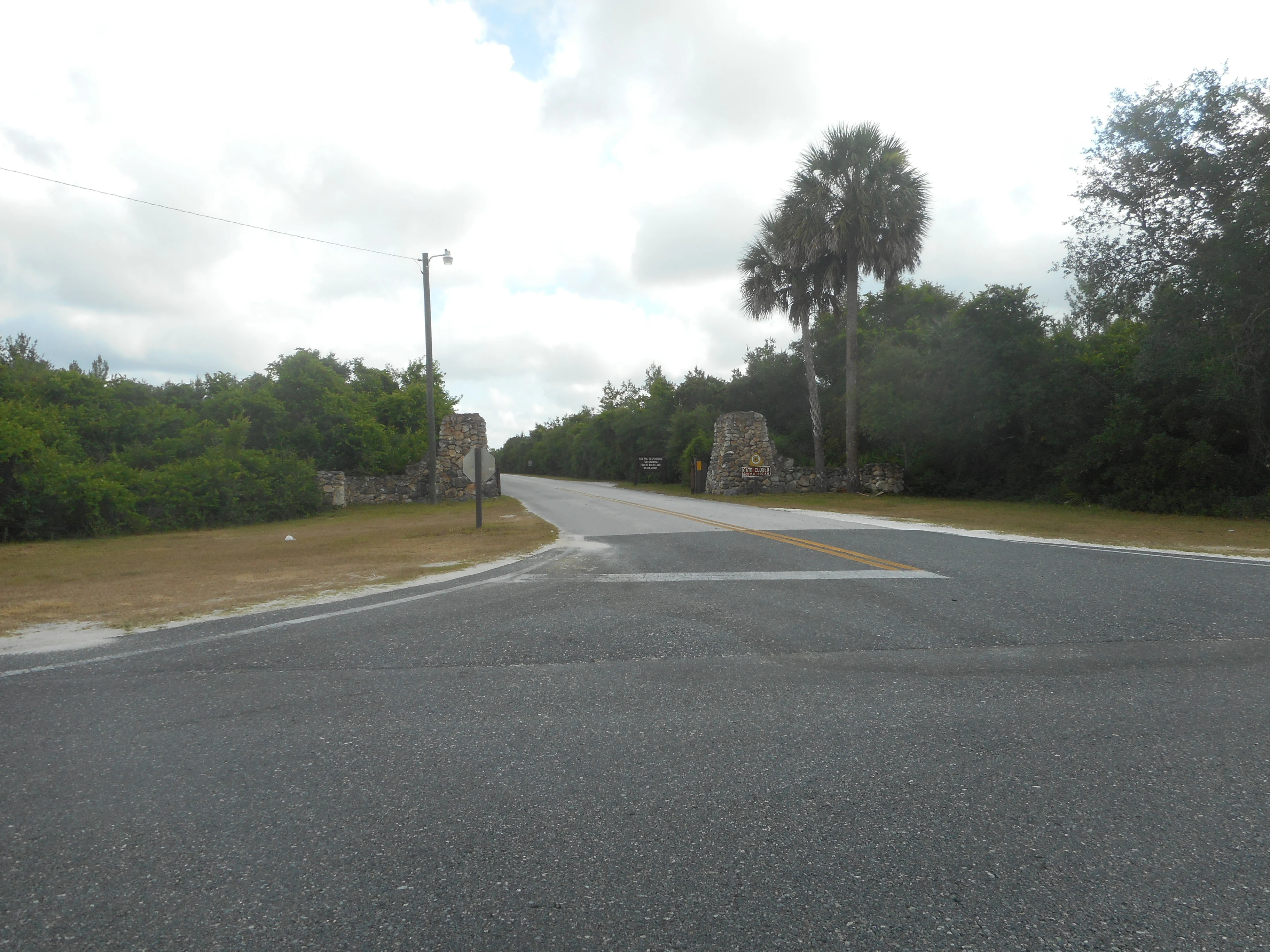 A paved road leading through a stone pillar gateway into a wooded recreation area, with lush Florida vegetation and palm trees in the background.