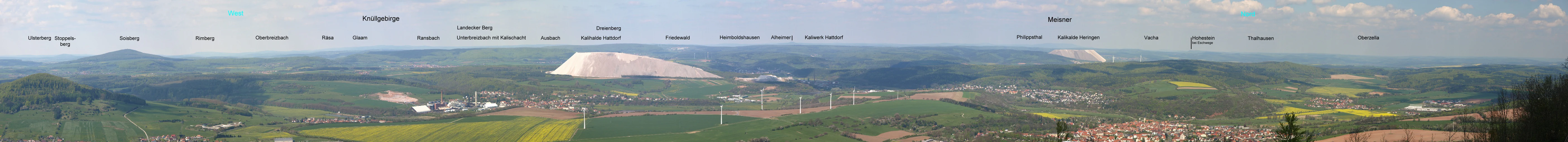 A wide panoramic view taken from a high vantage point, looking over a rolling green landscape with forests, agricultural fields, and a prominent white, cone-shaped industrial tailing heap in the distance.