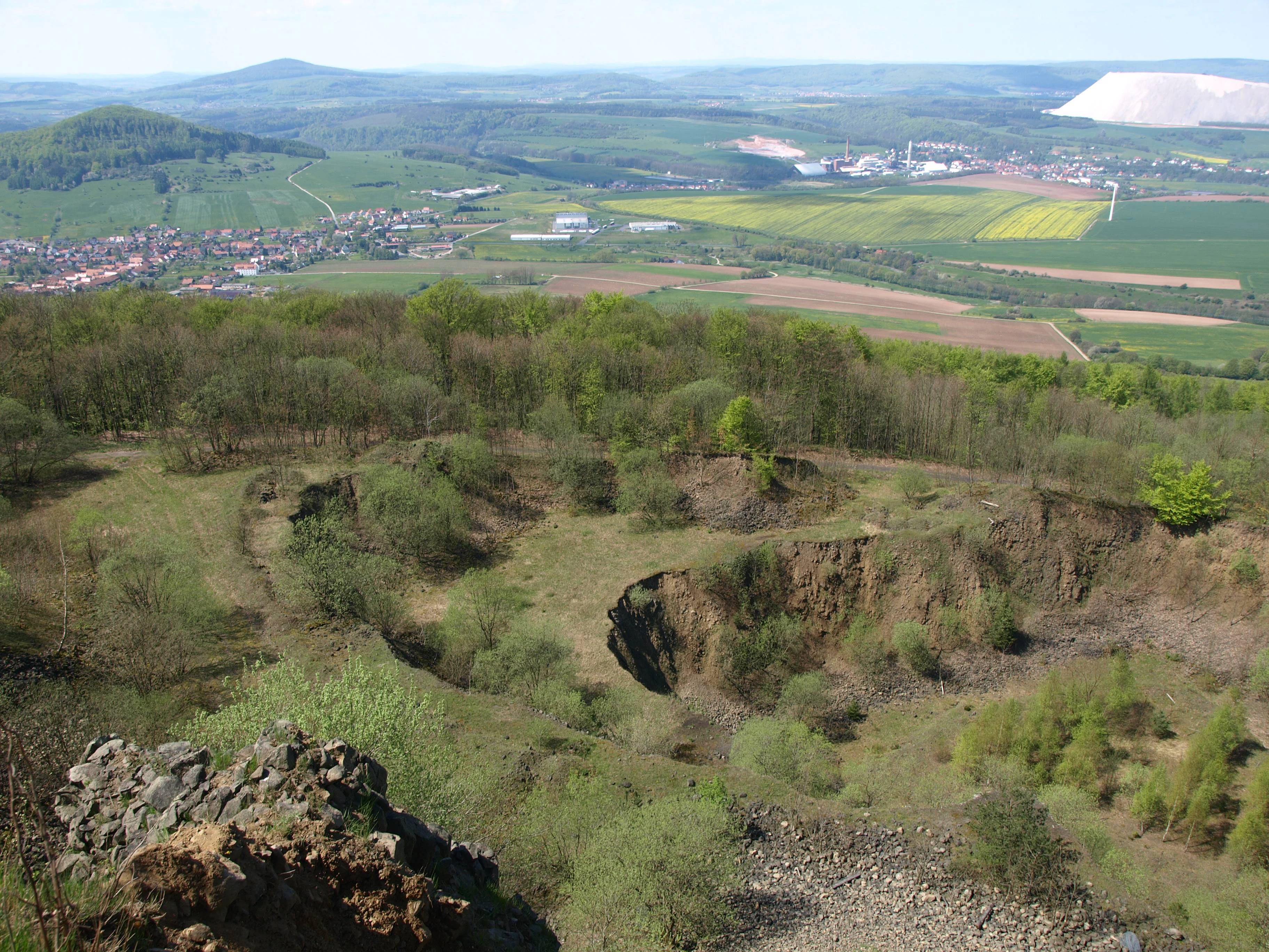 High angle shot of an abandoned, overgrown quarry with exposed rock faces and shrubs, looking down towards a green valley with a town, agricultural fields, and a distant white industrial slag pile.