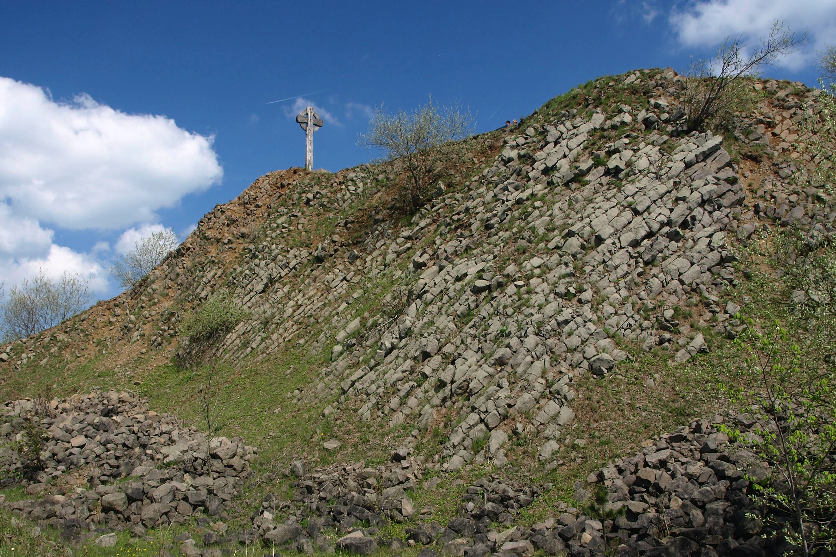 A sunny view of a steep, rocky basalt hillside with a wooden cross at the summit against a blue sky.