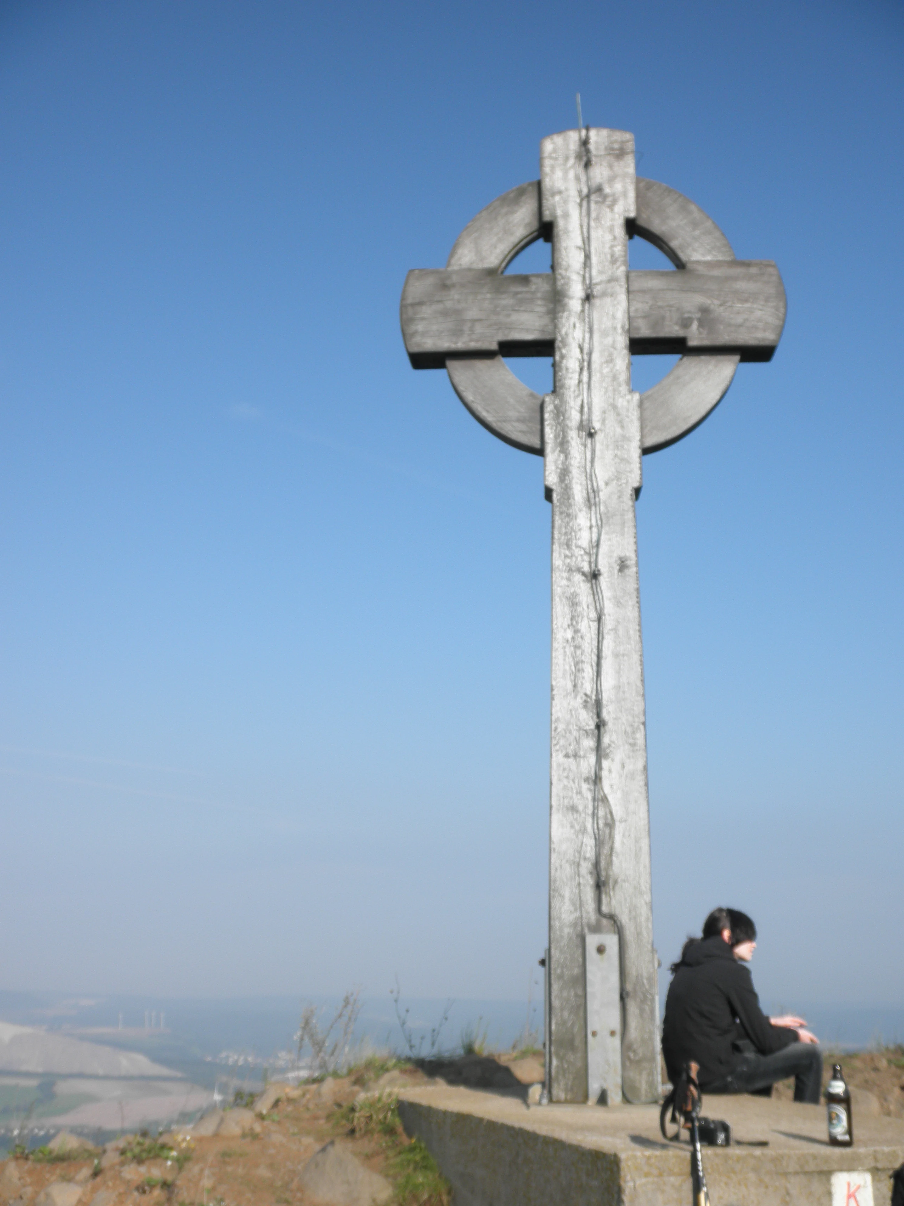 A large wooden Celtic cross stands on a concrete plinth at the top of a mountain, with a person sitting on the edge of the base looking out over the landscape.