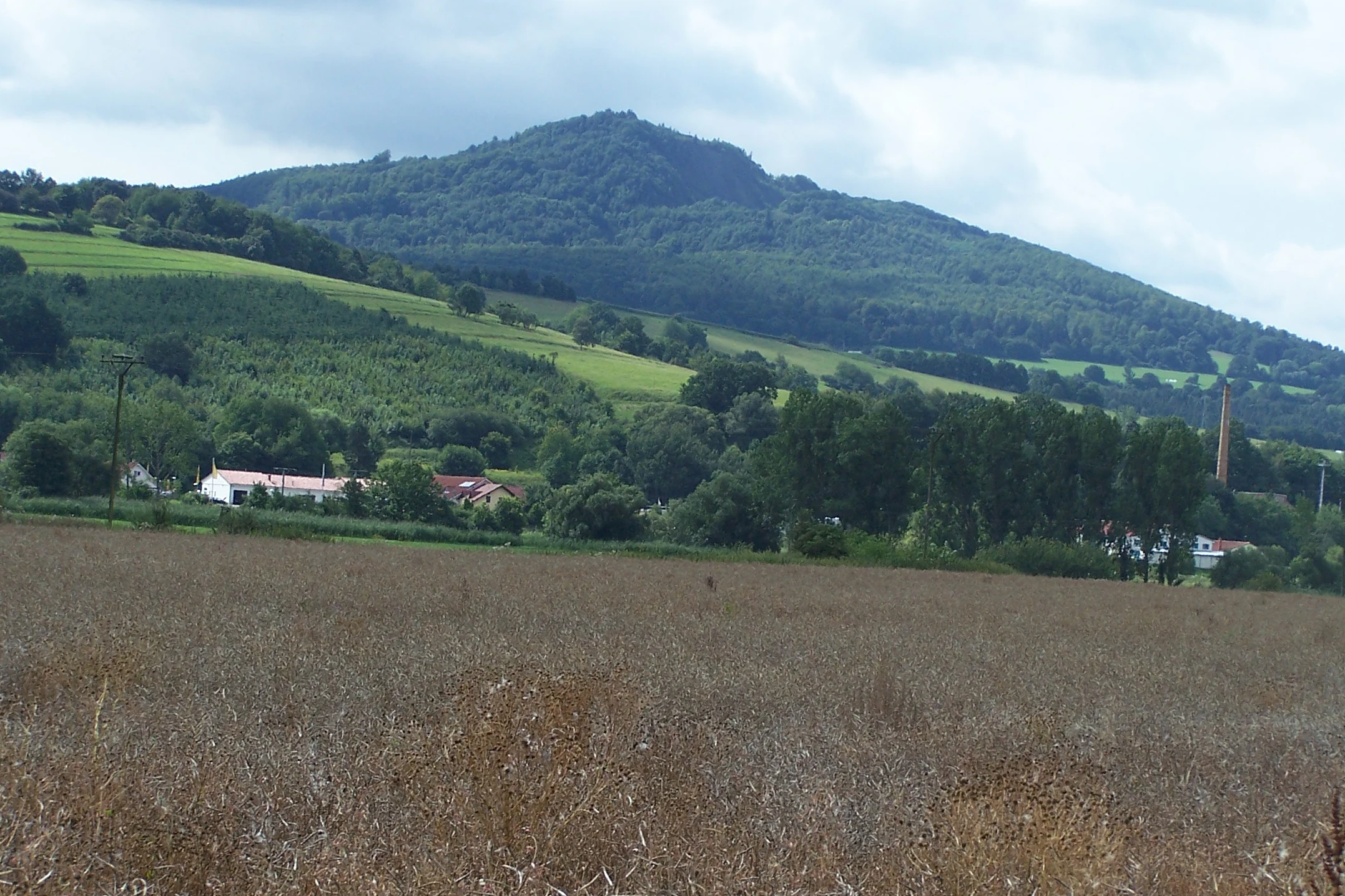 A rural landscape featuring a large, forested basalt mountain in the distance, viewed across an agricultural field with a few small buildings and a chimney visible at the base.