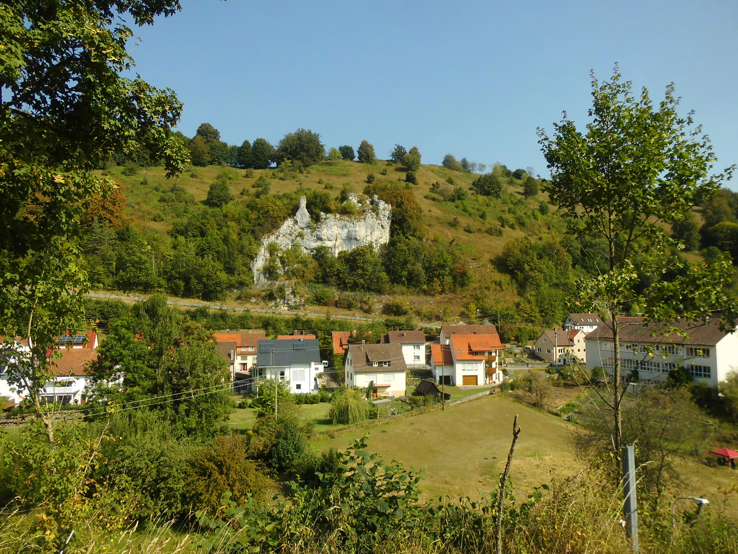 A sunny day view of a light-colored limestone rock cliff emerging from a grassy and wooded hillside, with residential houses of a small village visible at the base of the slope.