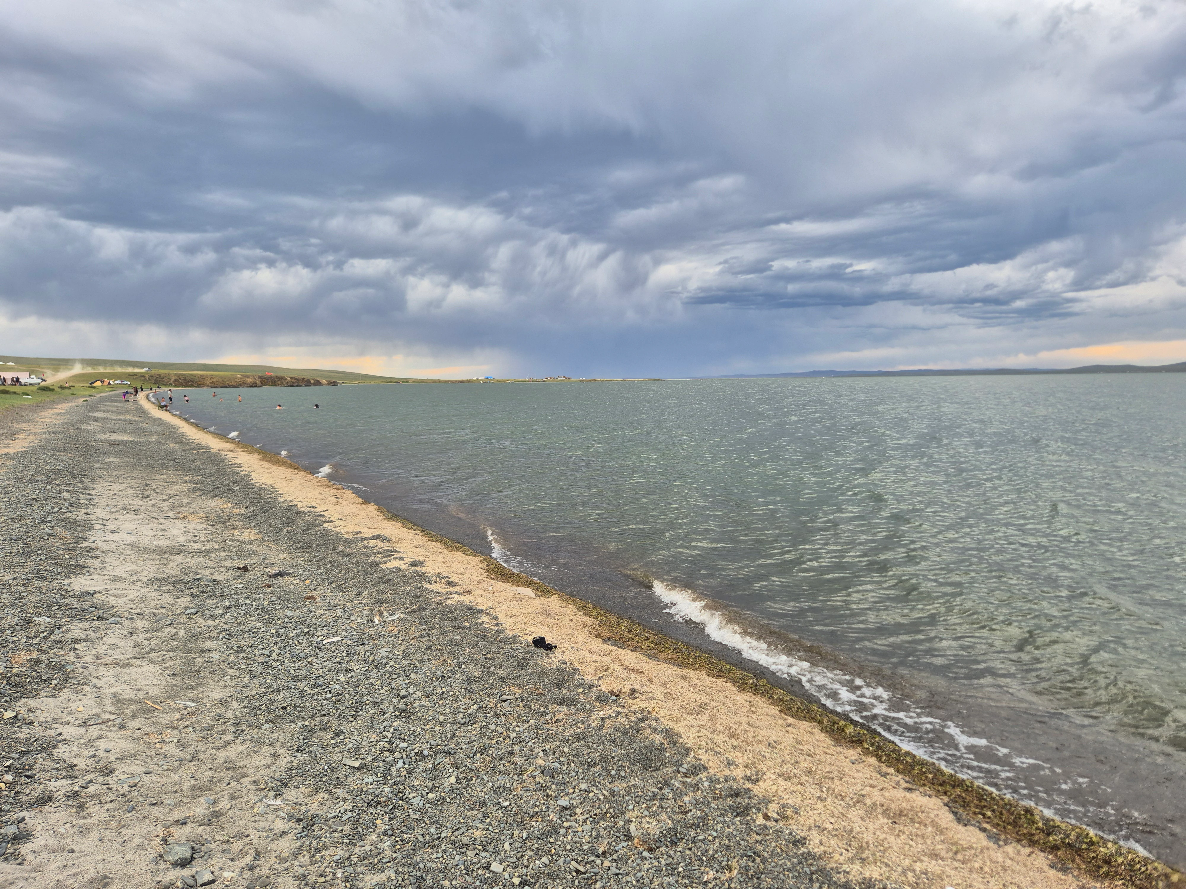 A wide, low-angle shot of the gravelly shoreline of a large, calm lake under a heavy, grey, cloudy sky in Mongolia.