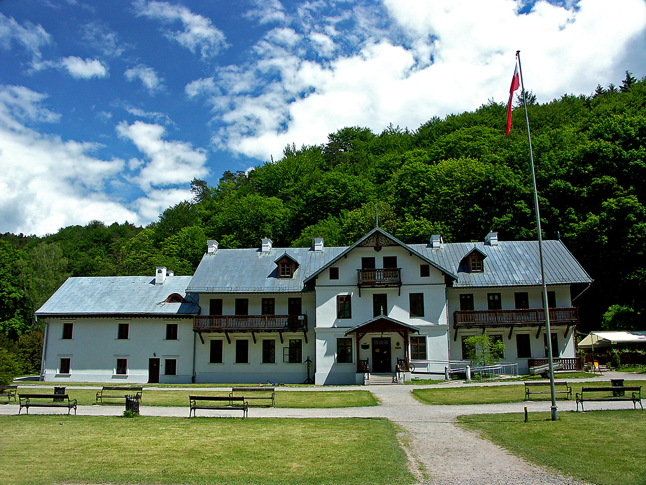 White building with gray roof, flagpole with red flag, grassy lawn, benches, and forested hill