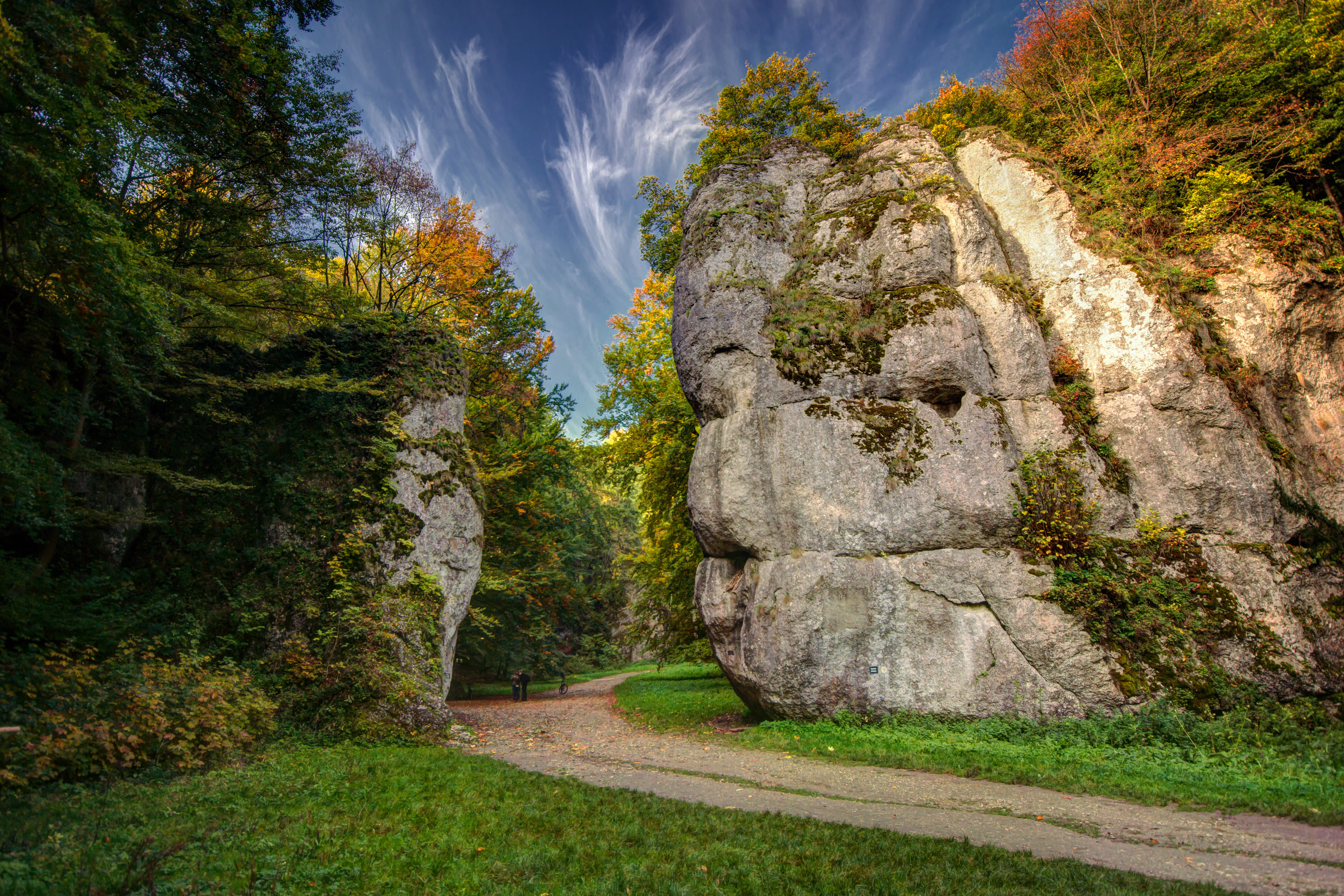 Large limestone rock formation with vegetation, a dirt path, and forested area under a partly cloudy sky