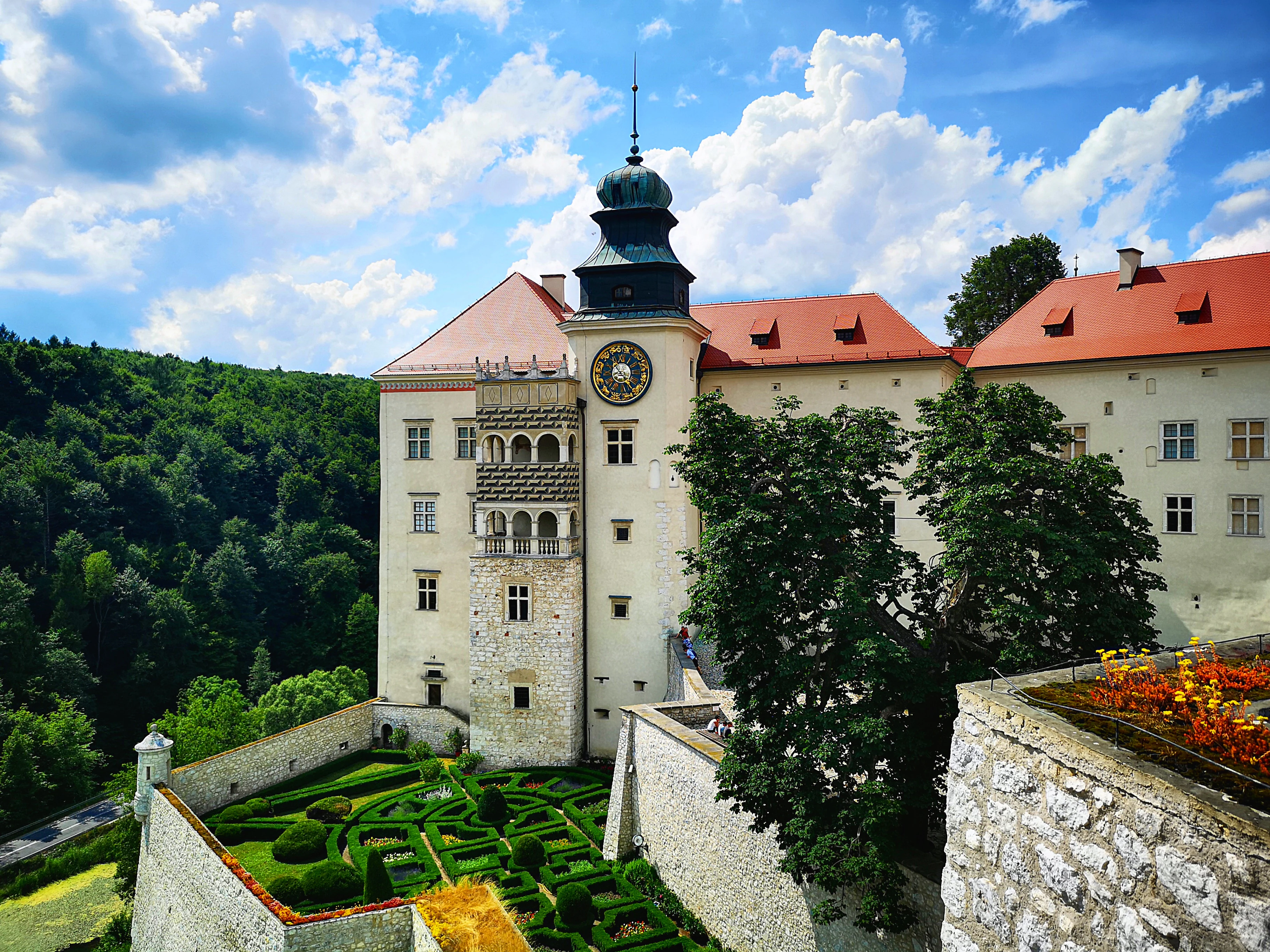 A historic castle with red roofs and a clock tower, surrounded by stone walls and manicured gardens under a partly cloudy sky