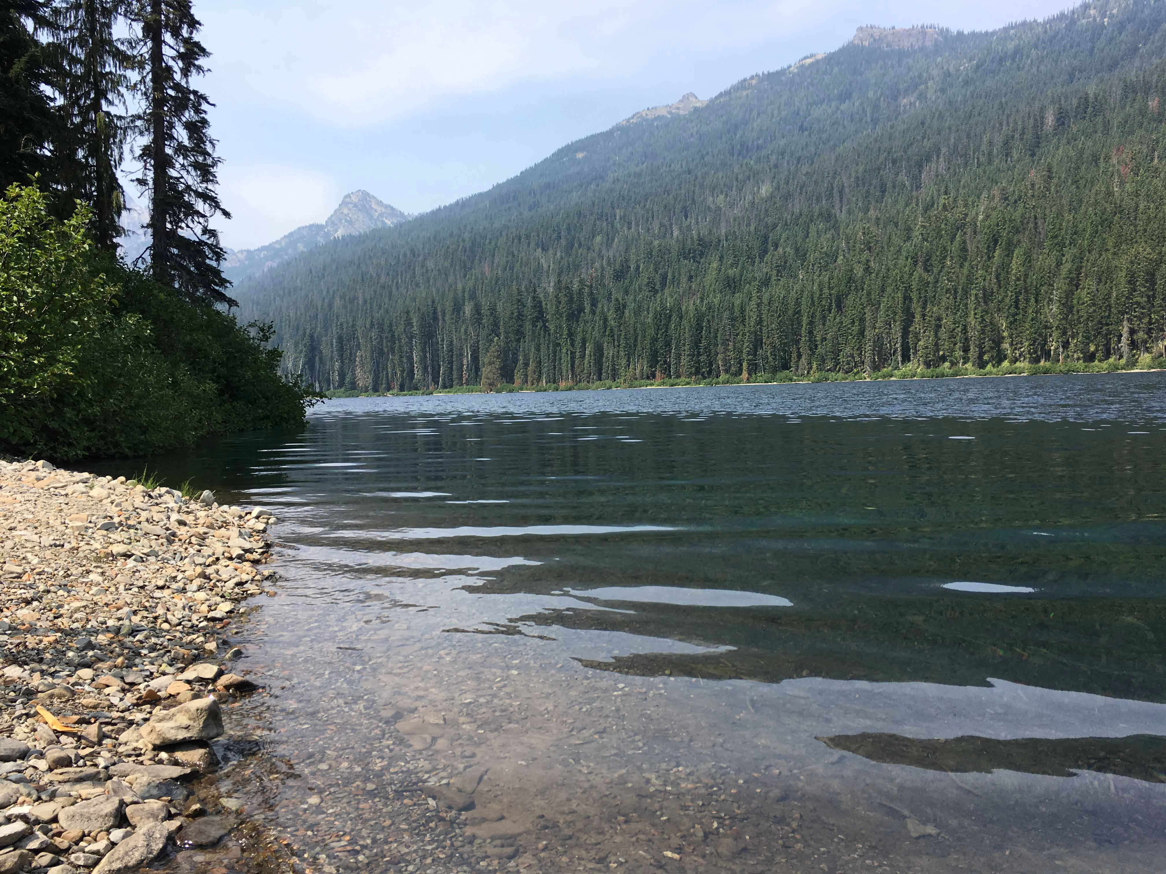 A rocky shoreline borders a calm lake, with dense evergreen forest covering the slopes of a mountain in the background under a partly cloudy sky.