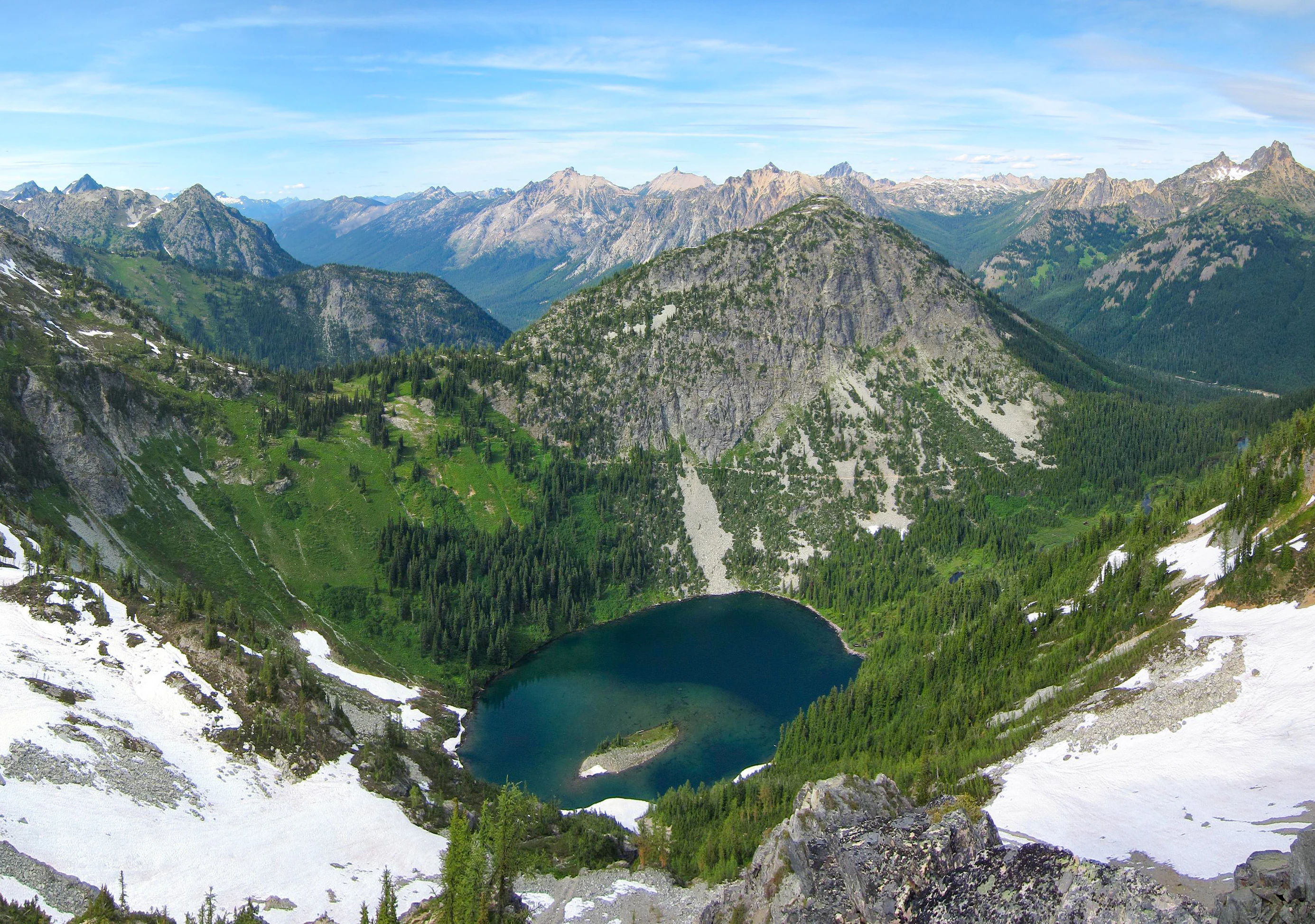 A high angle landscape photograph showing a deep blue alpine lake surrounded by green coniferous forests and rocky mountain peaks, with patches of snow in the foreground under a blue sky.