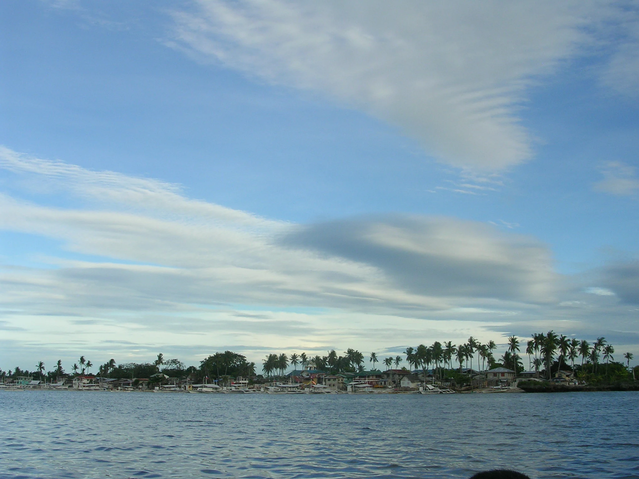 A wide view of a low-lying island covered in palm trees and scattered buildings, seen from across calm, open water under a blue sky with streaky clouds.