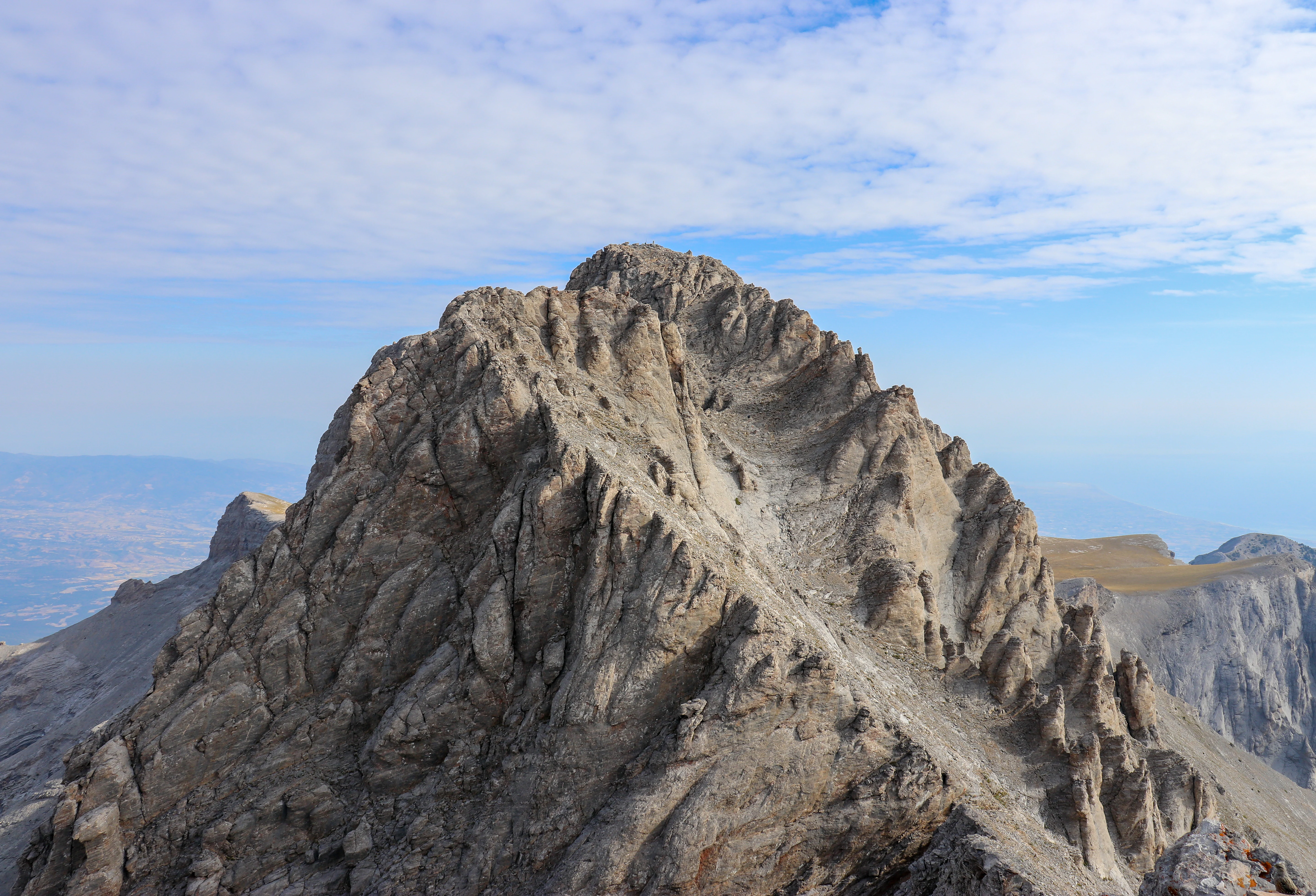 A high-angle view of the sharp, rocky summit of Mytikas peak on Mount Olympus, showing textured stone faces and distant hazy mountain ranges under a partly cloudy sky.