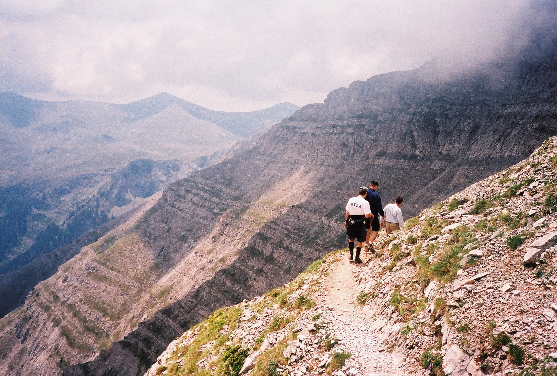 A group of three hikers walking along a narrow, rocky mountain trail with a steep drop-off on the left and a dramatic mountainous landscape in the background.