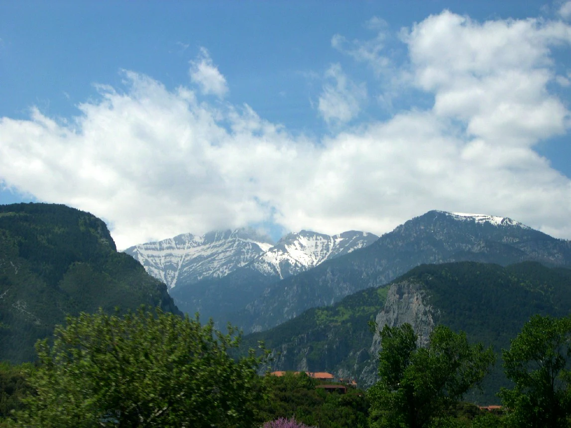 A wide view of Mount Olympus with snow-covered peaks under a partly cloudy blue sky, framed by forested slopes in the foreground.